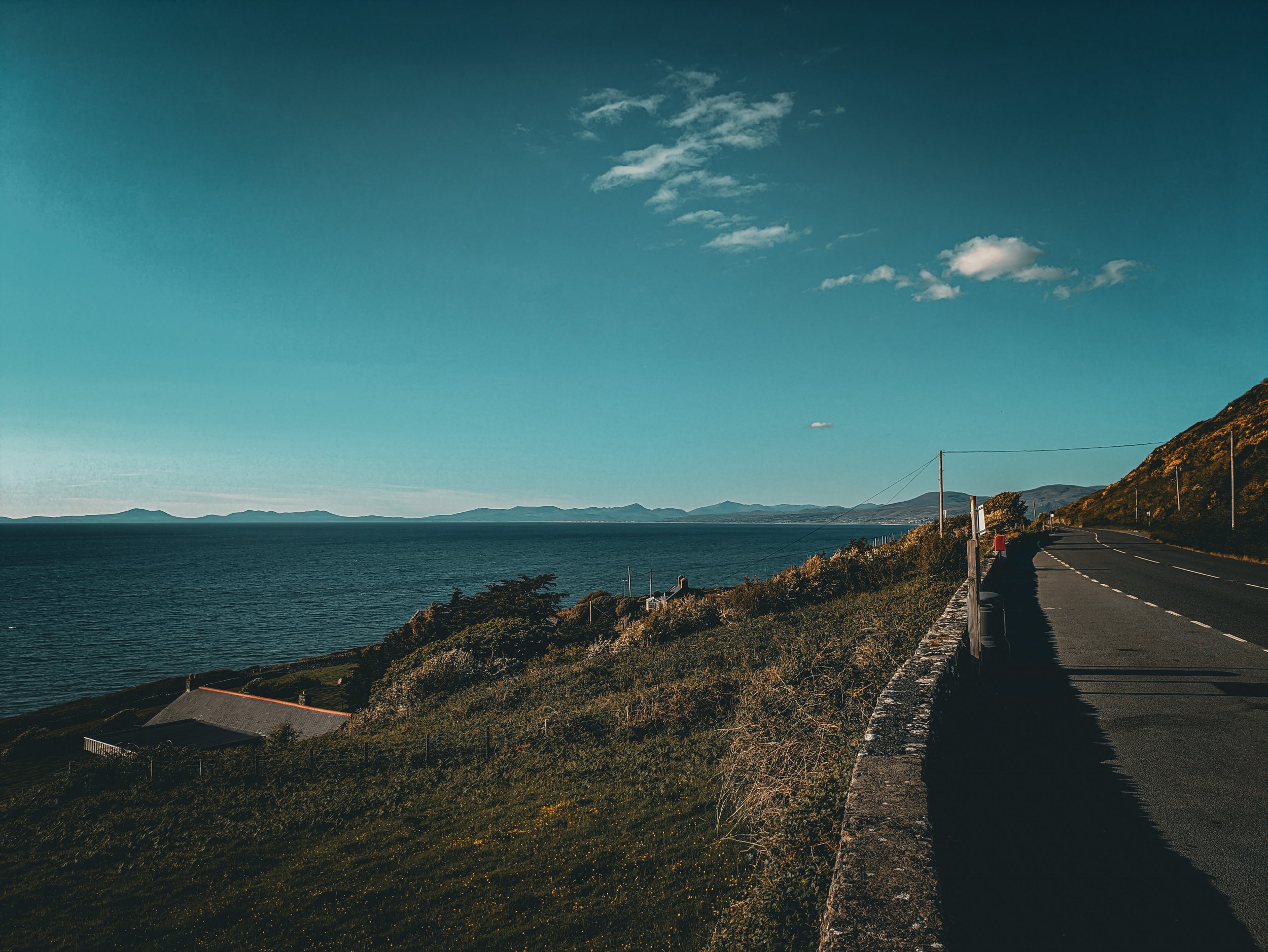 A coastal road runs along the base of a hillside with houses and vegetation, overlooking a large body of water with mountains in the distance under a blue sky with some clouds.