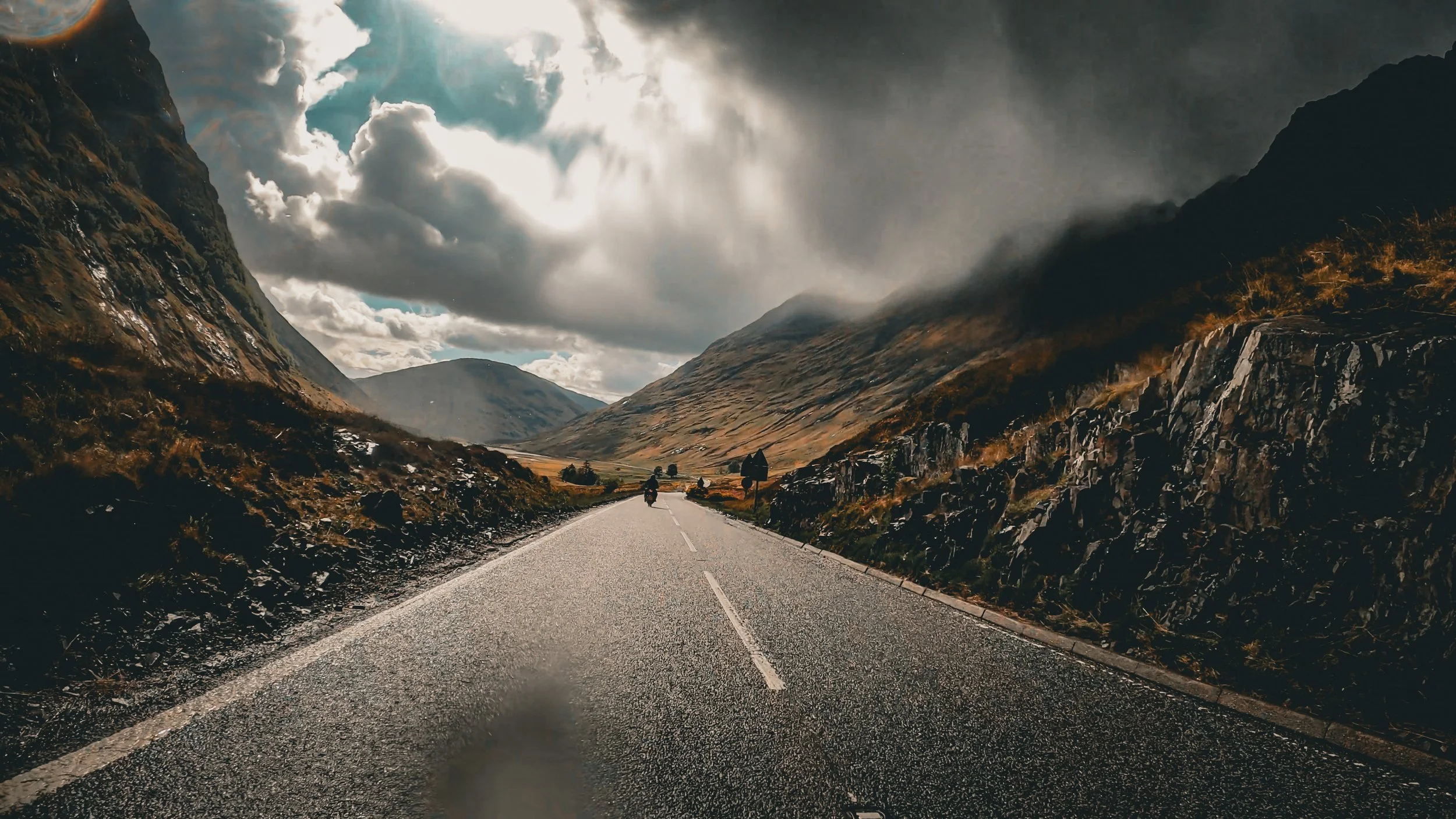 A motorcycle on a desolate highway surrounded by towering mountains with dark clouds overhead, creating a dramatic landscape.