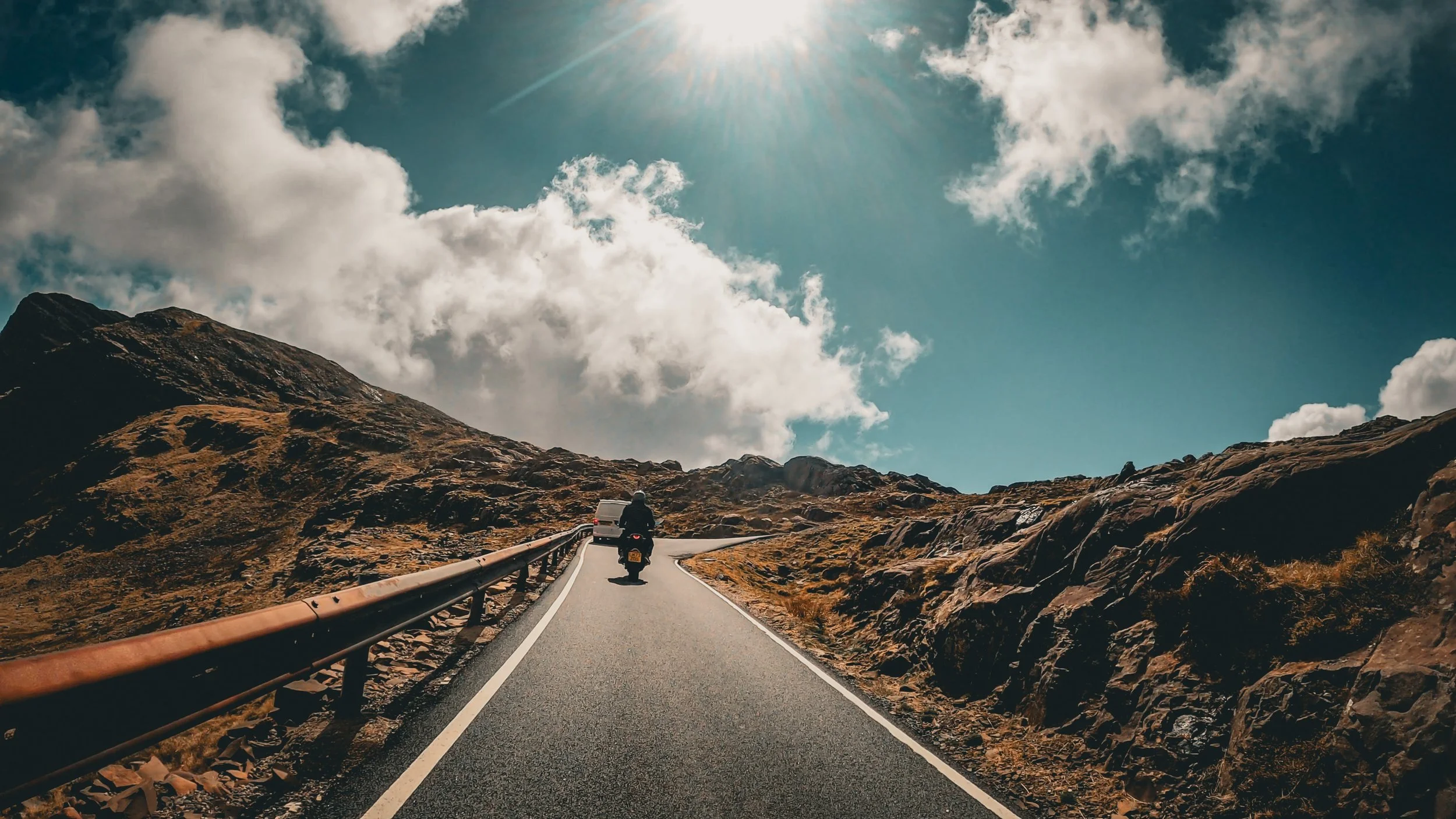 Motorcycle rider traveling on a winding mountain road under a partly cloudy sky with sunlight. Rocky terrain on either side of the road.