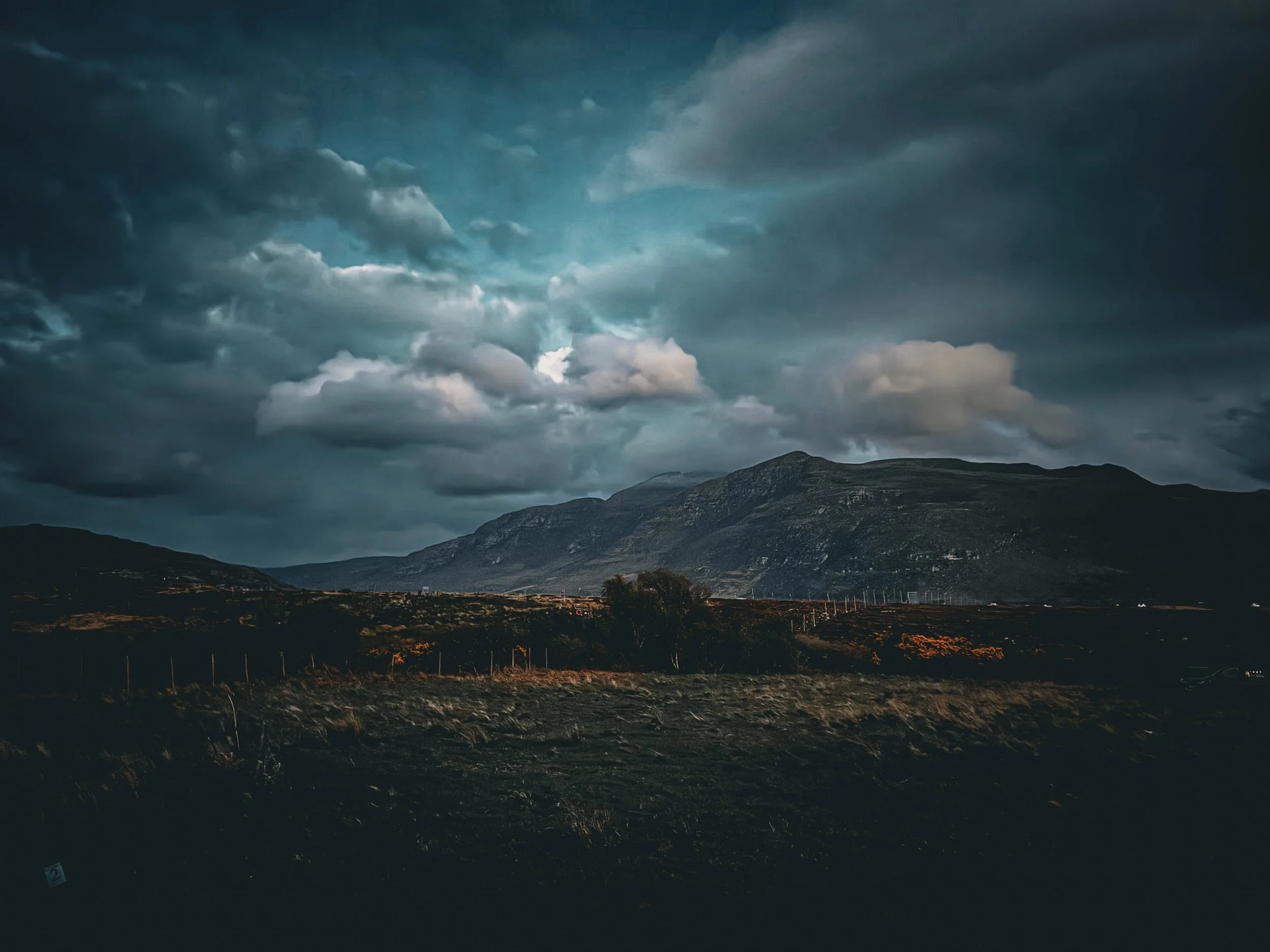 Dark clouded sky over a mountainous landscape with a fence and some trees in the foreground during twilight.
