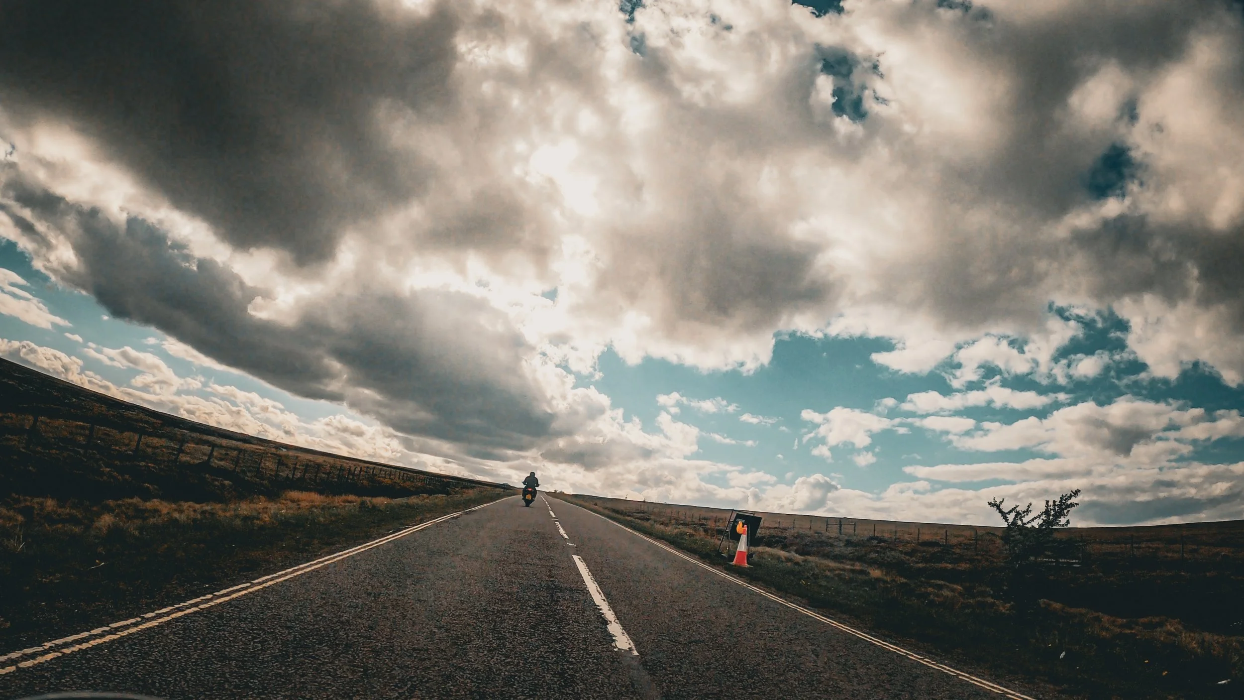 A motorcyclist riding along an open road under a cloudy sky, with a traffic cone on the side of the road.