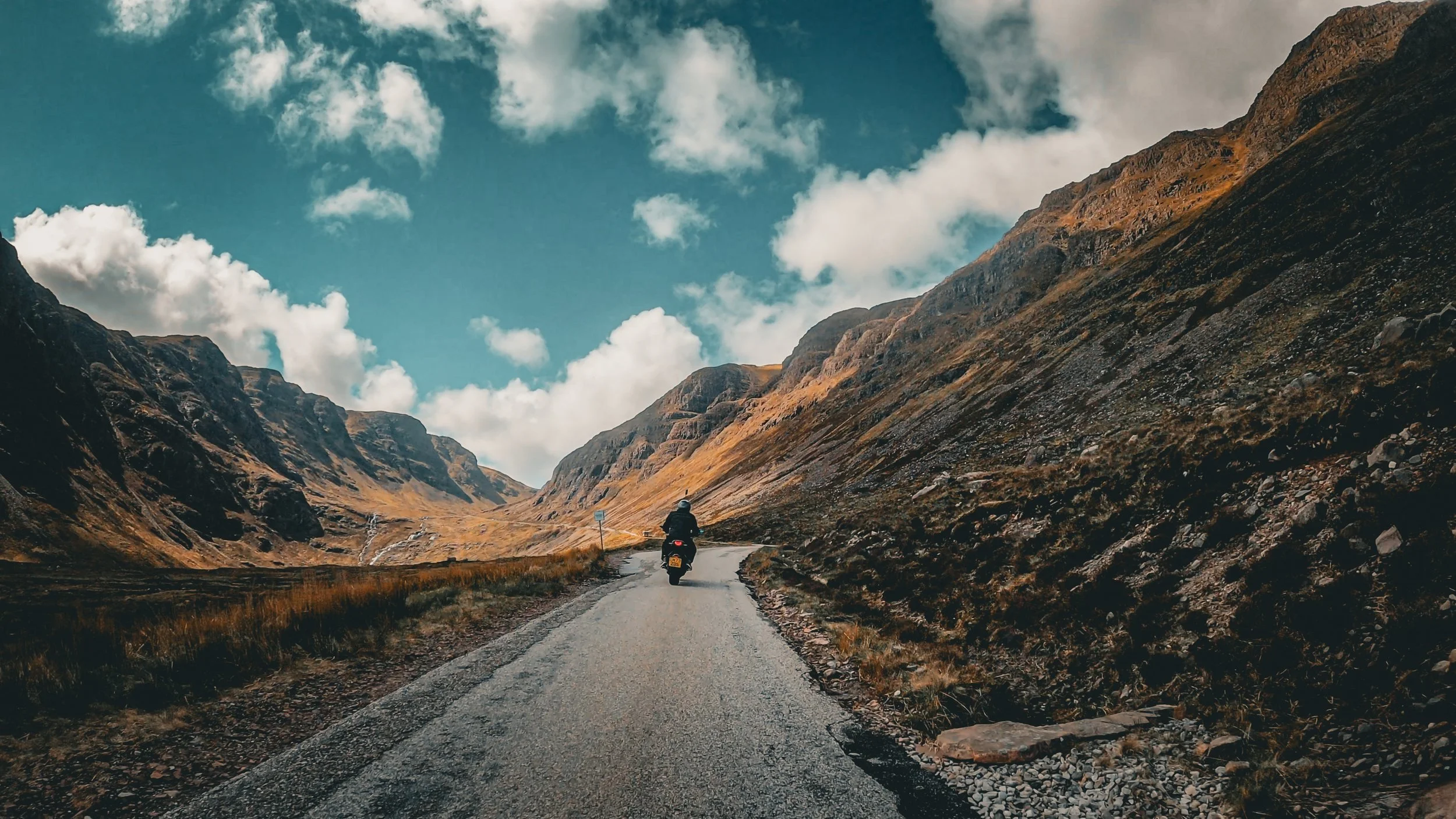 A person riding a motorcycle on a winding mountain road with tall, rugged hills on both sides and a partly cloudy sky overhead.
