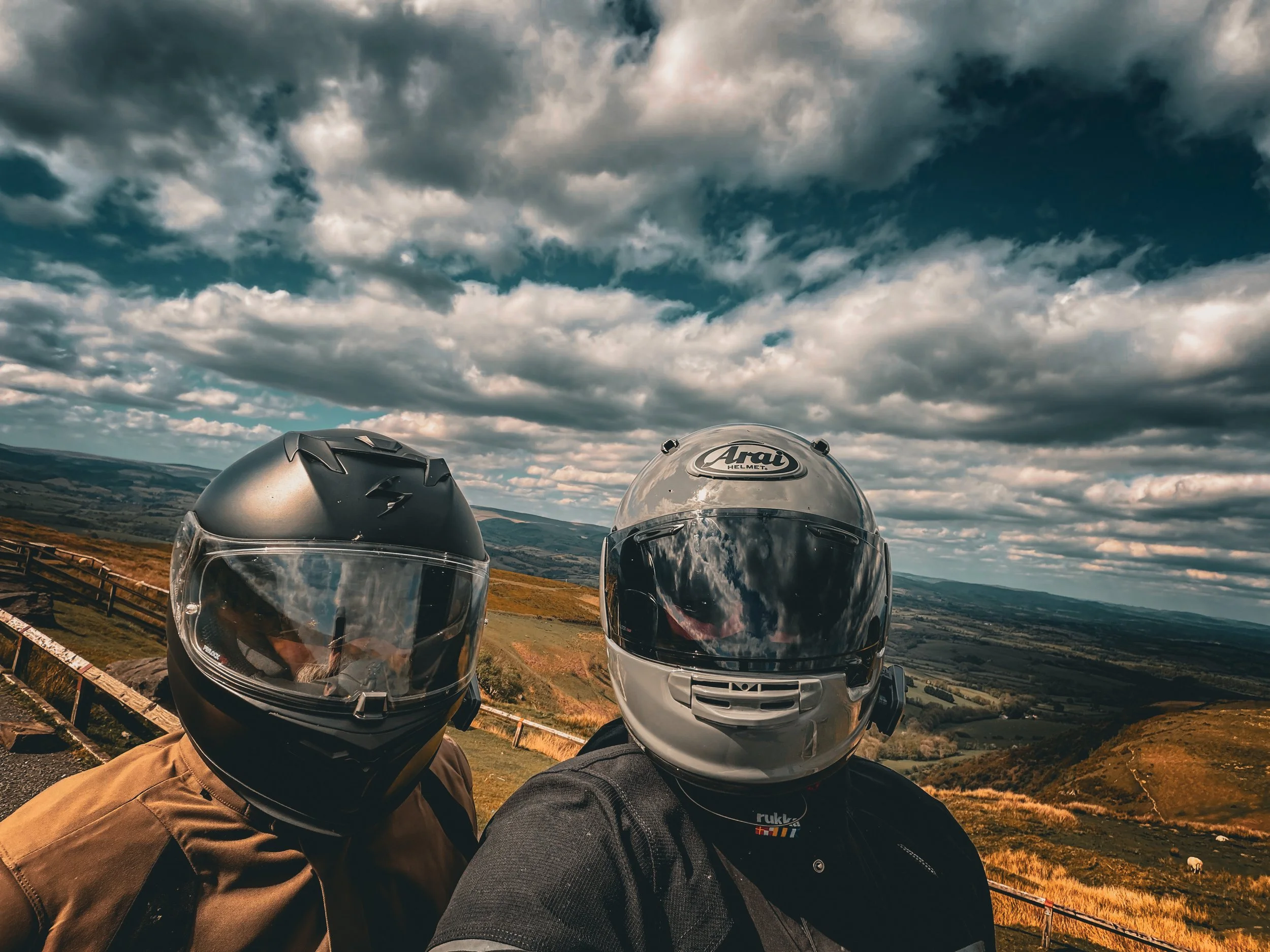 Two motorcyclists wearing helmets and jackets taking a selfie outdoors in a hilly landscape with cloudy skies.