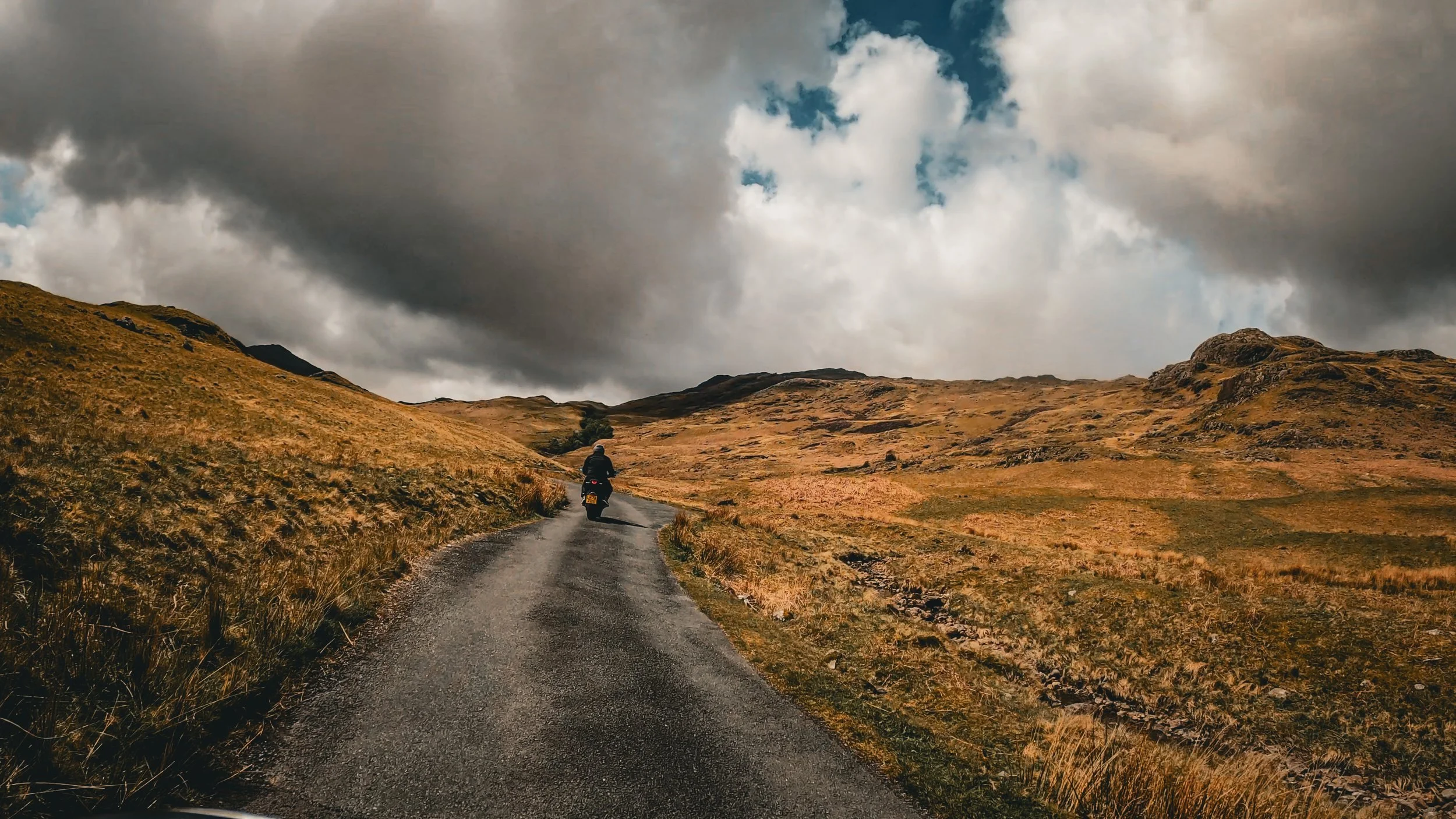A person riding a motorcycle on a narrow country road through a barren, hilly landscape with cloudy sky overhead.