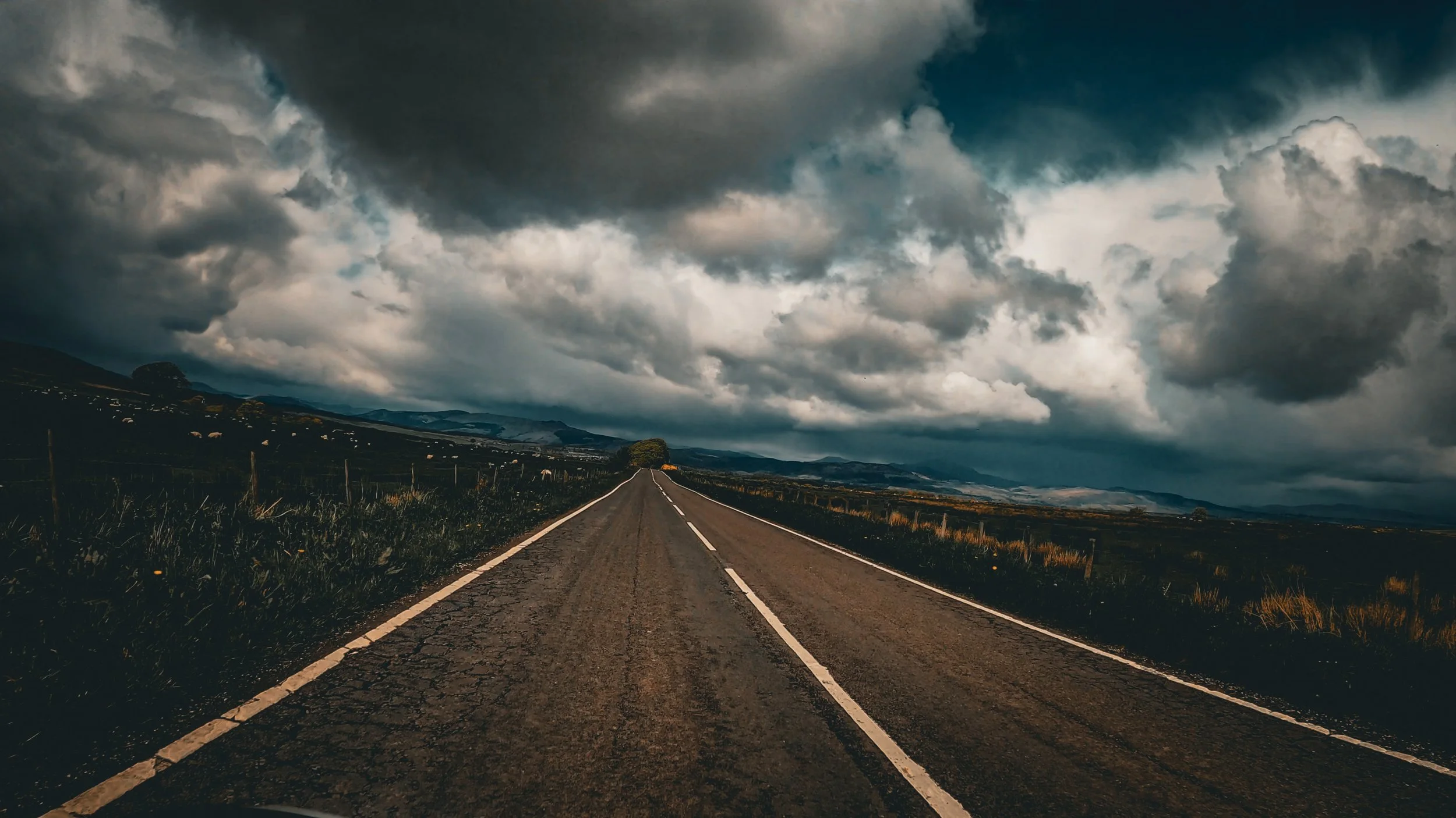 A long empty road stretching into the distance under a dark, cloudy sky, flanked by fields and hills.