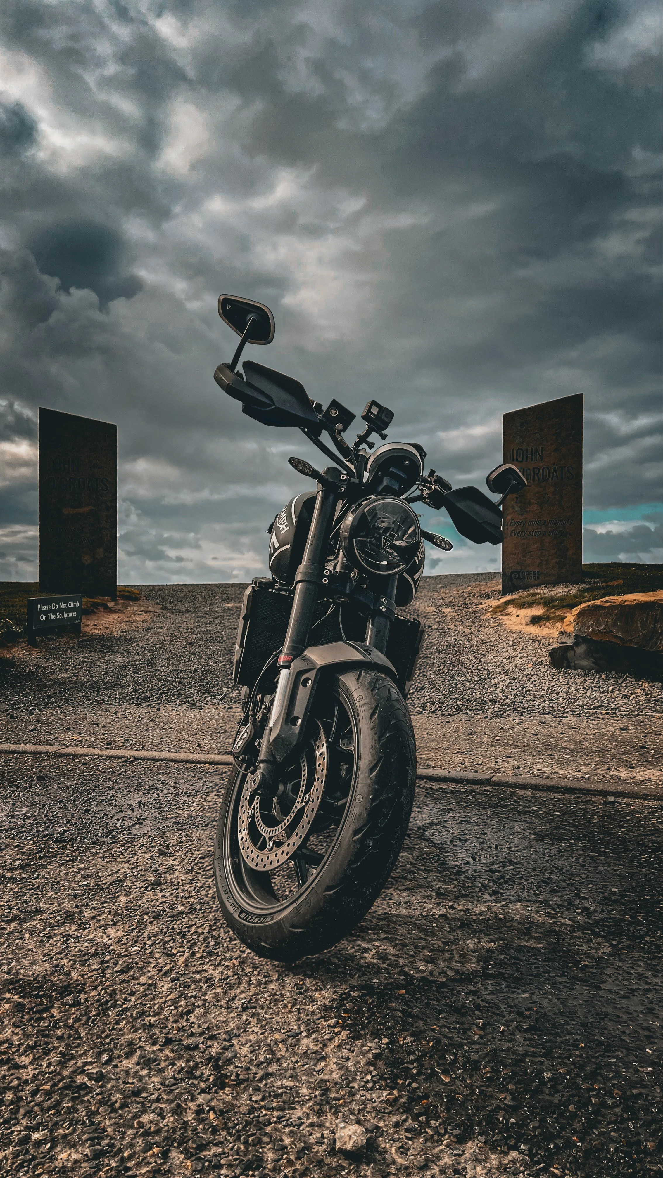 A black motorcycle parked on a gravel path under a cloudy sky with two large signs and a small sign that says "Please Do Not Climb On The Sculptures."