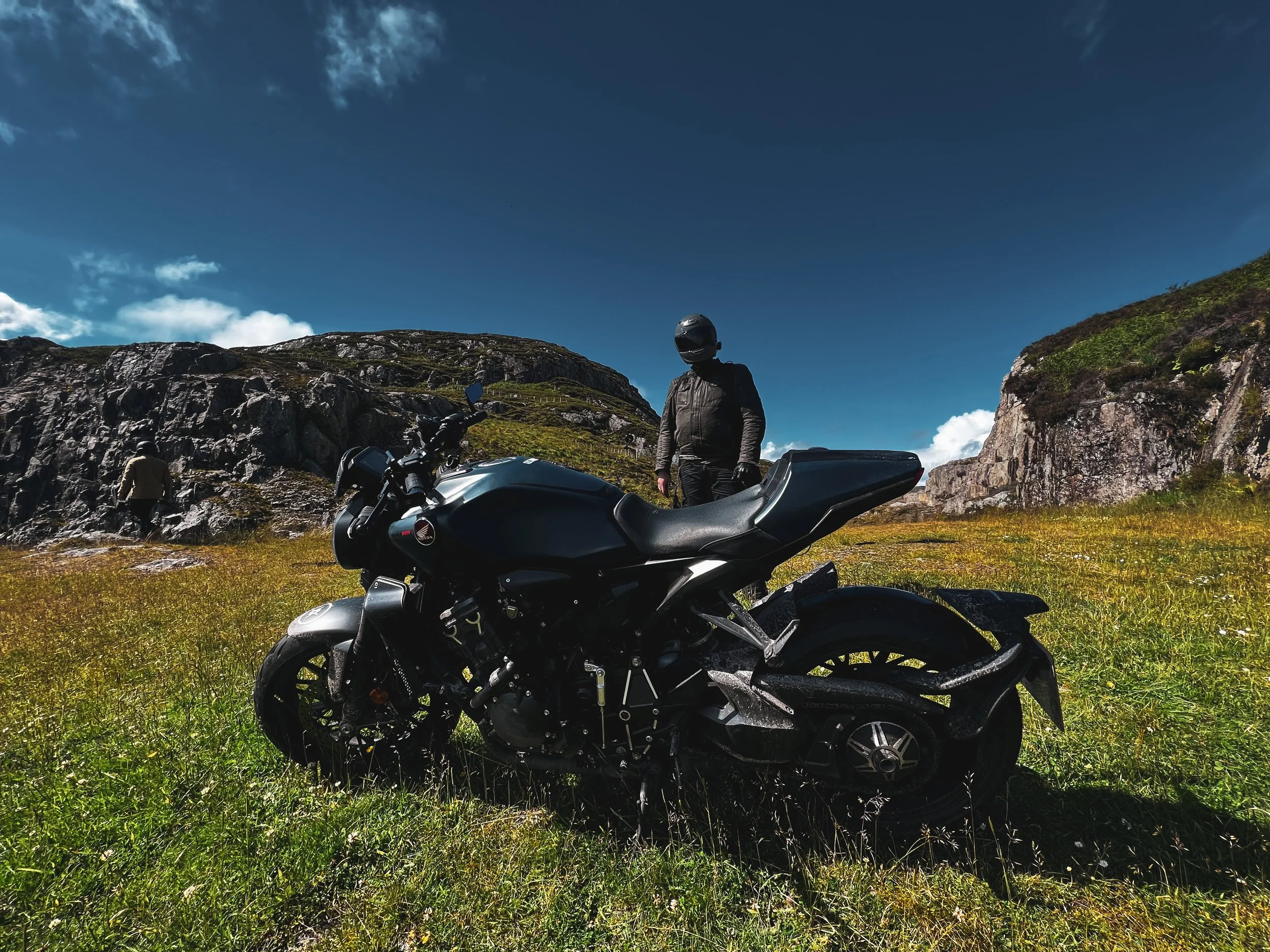 Motorcycle parked on grassy terrain in front of rocky hills with two people nearby, with a person standing and another walking on a sunny day with a clear sky.