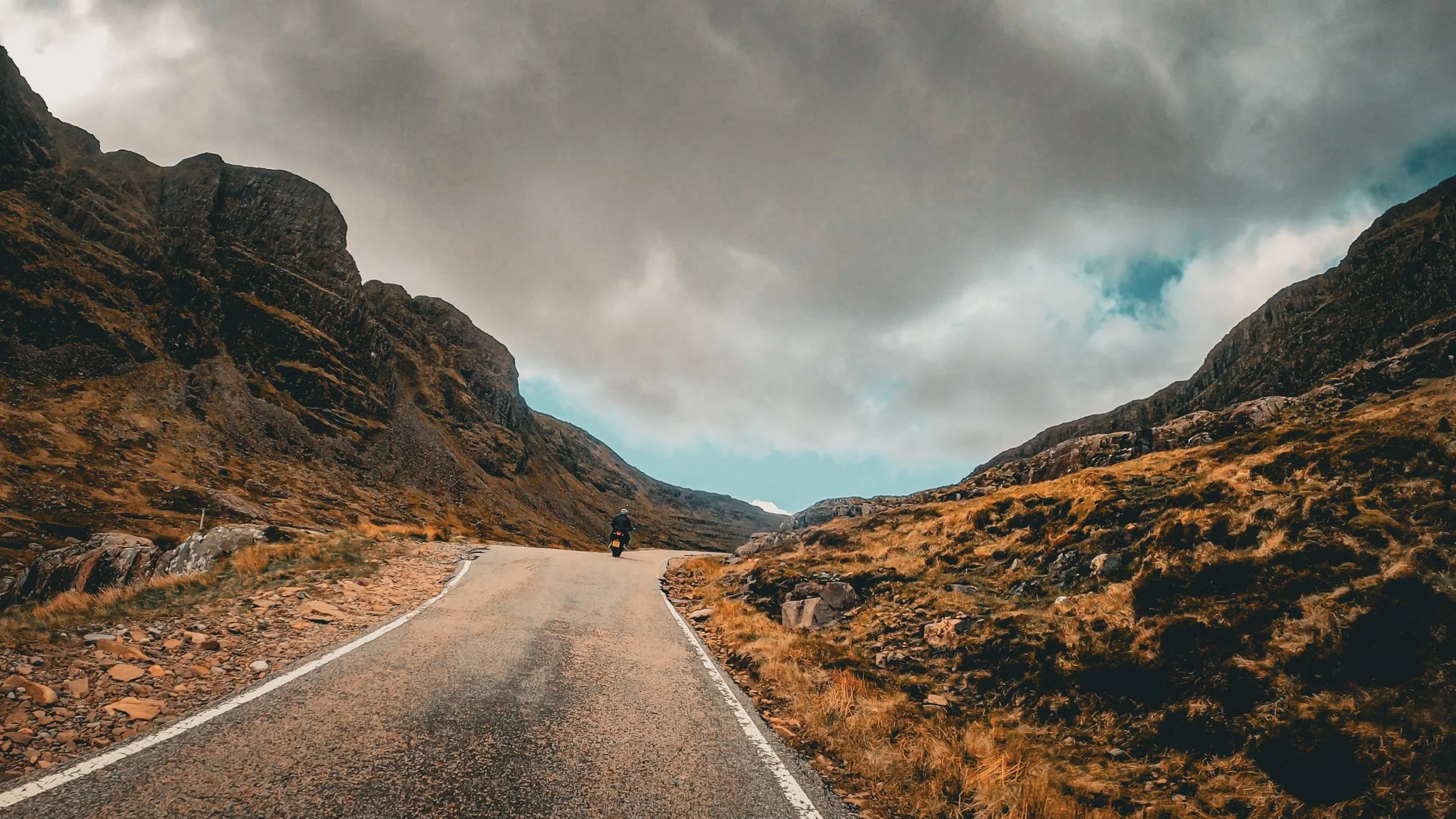 Motorcyclist riding on a winding mountain road surrounded by rocky hills and cloudy sky.