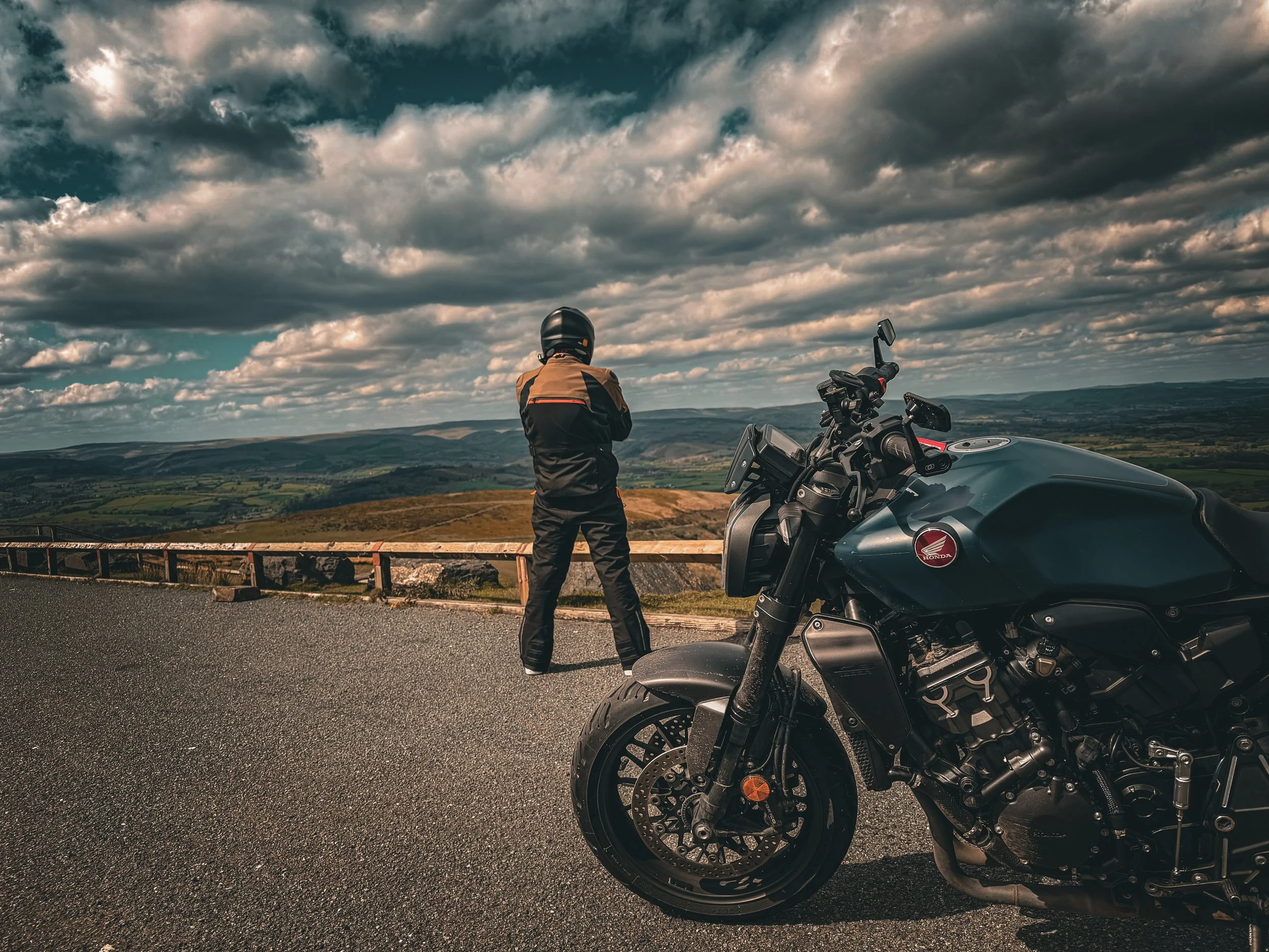 A person wearing motorcycle riding gear stands next to a parked black motorcycle with a Honda logo, overlooking a panoramic landscape with rolling hills and a cloudy sky.