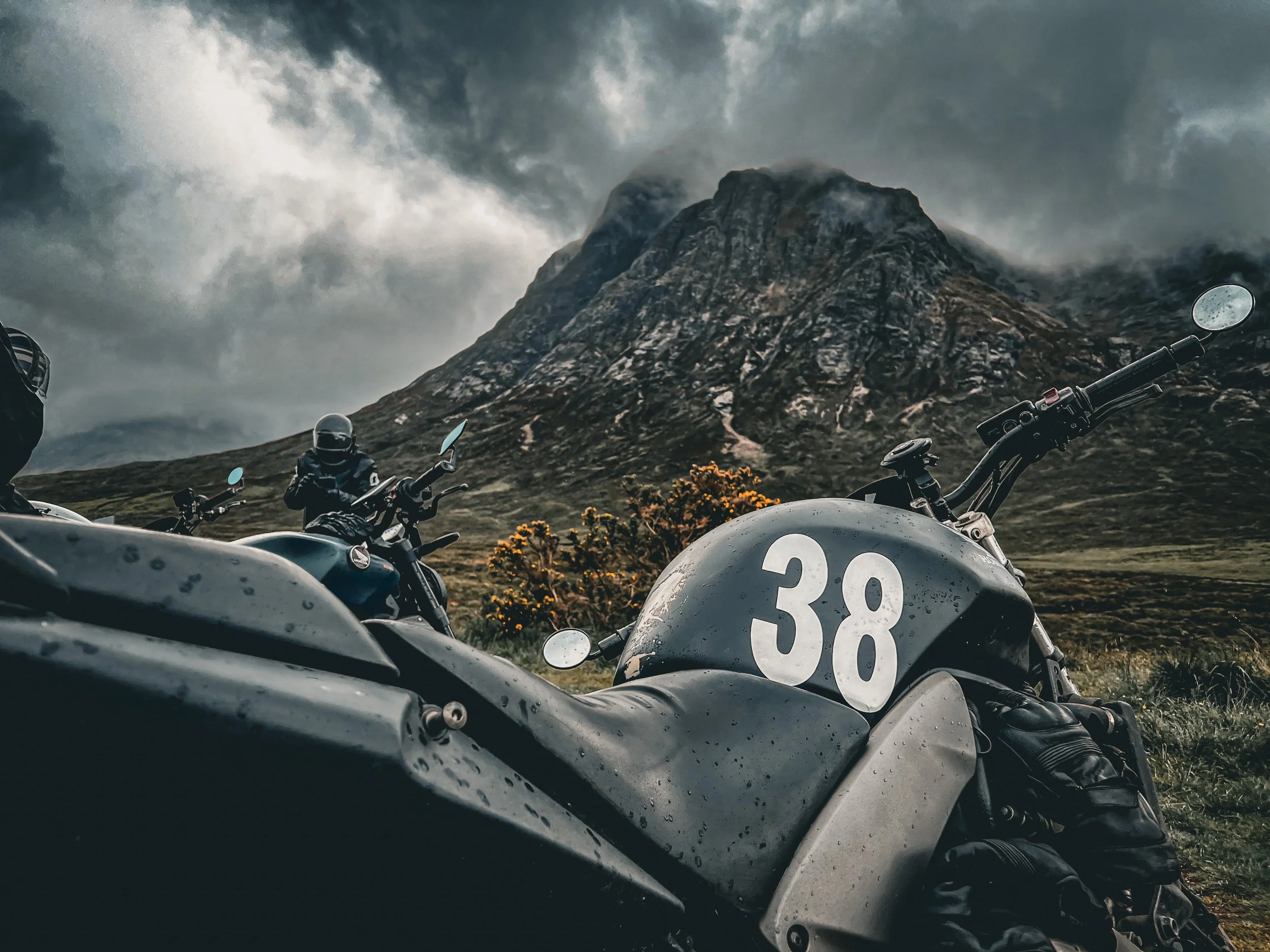 Motorcycles parked on a grassy hillside with rugged mountain peak and cloudy sky in the background.