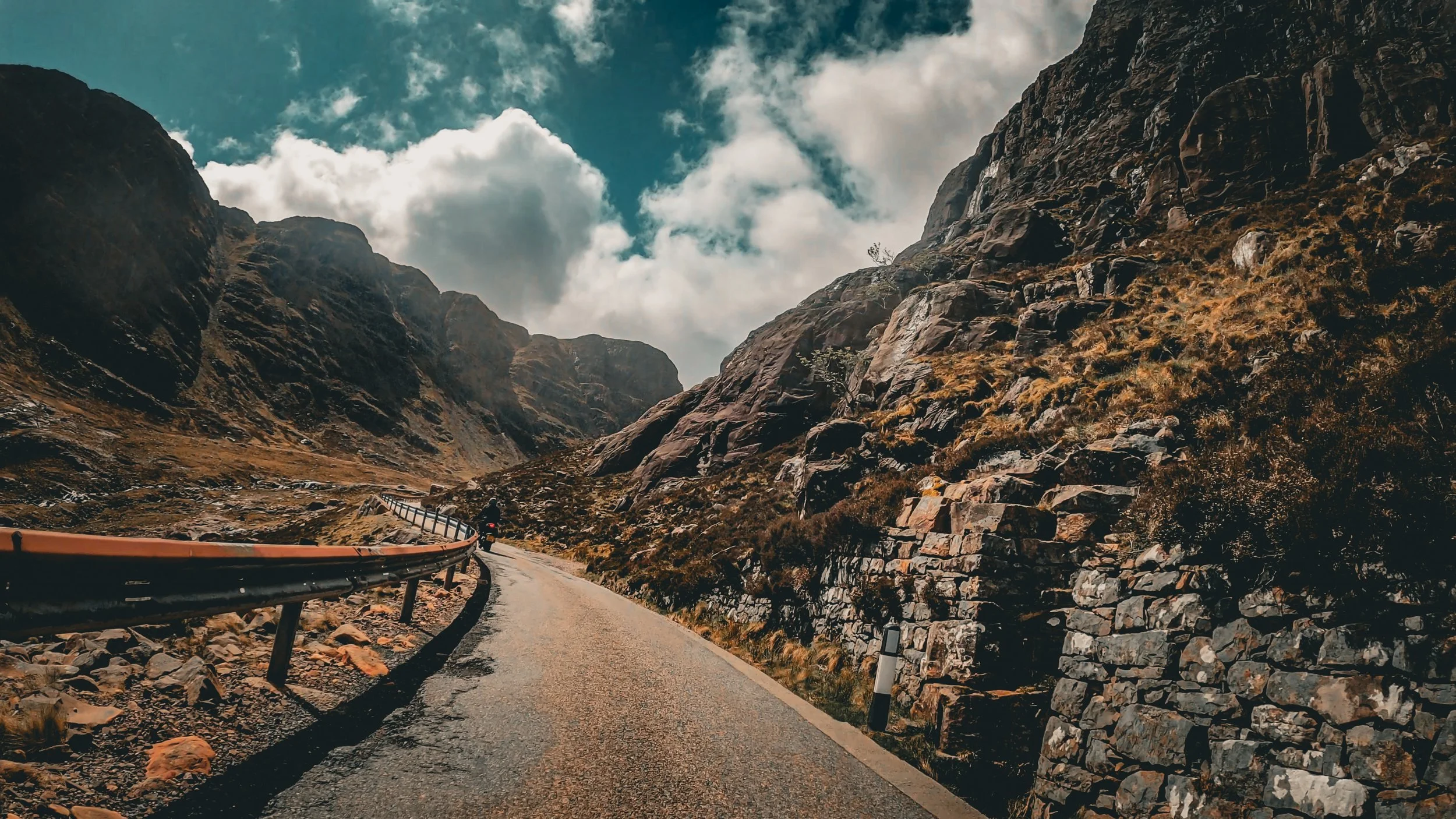 A winding mountain road with a guardrail on one side, surrounded by rocky cliffs and steep slopes under a partly cloudy sky.