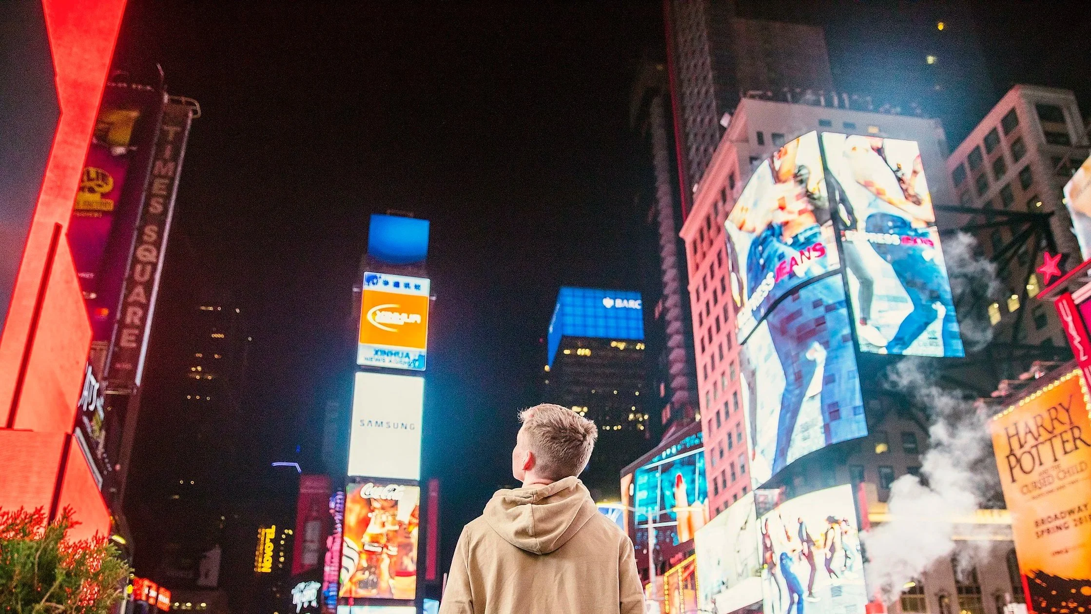 A person with short blond hair and tan jacket standing in Times Square at night, surrounded by bright electronic billboards and advertisements, with one person taking a photo.
