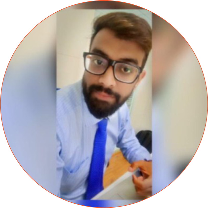 A young man with glasses and a beard, wearing a business shirt and blue tie, sitting at a desk in an office environment.