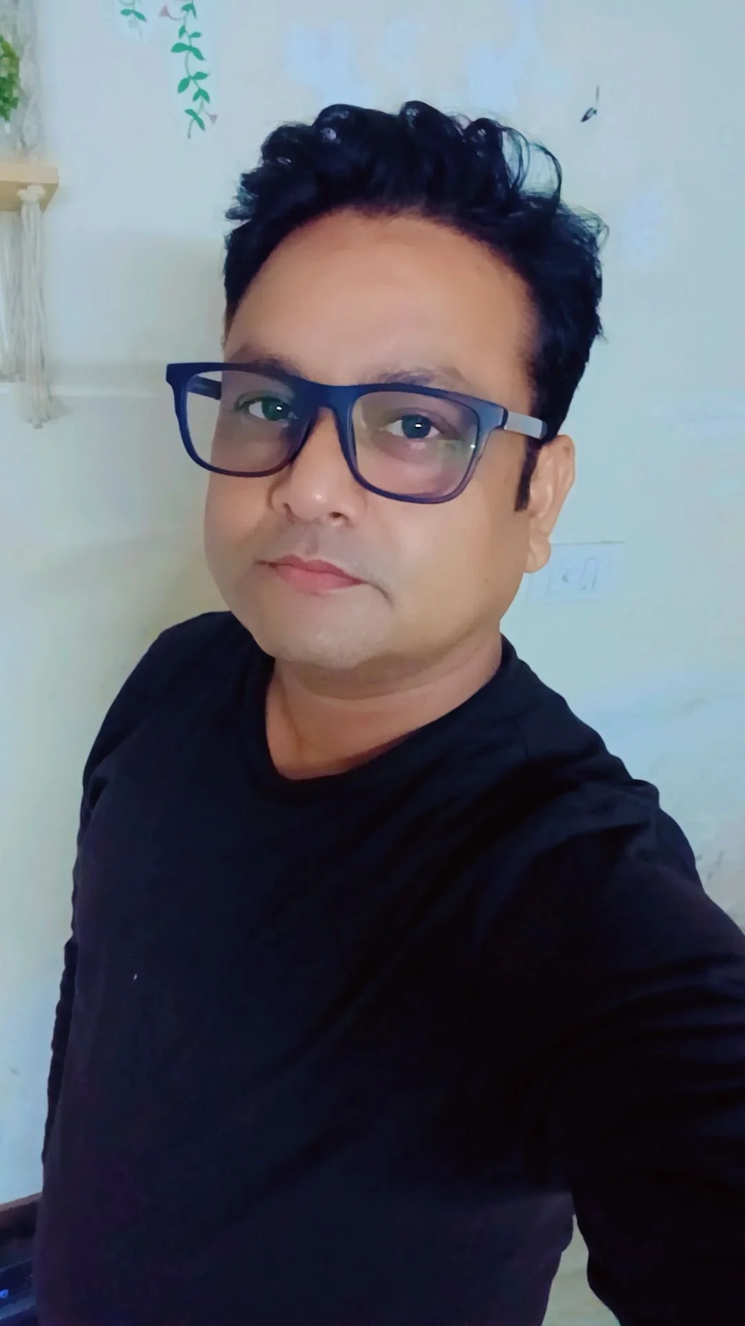 A man with short, curly black hair and wearing glasses and a black shirt taking a selfie indoors.