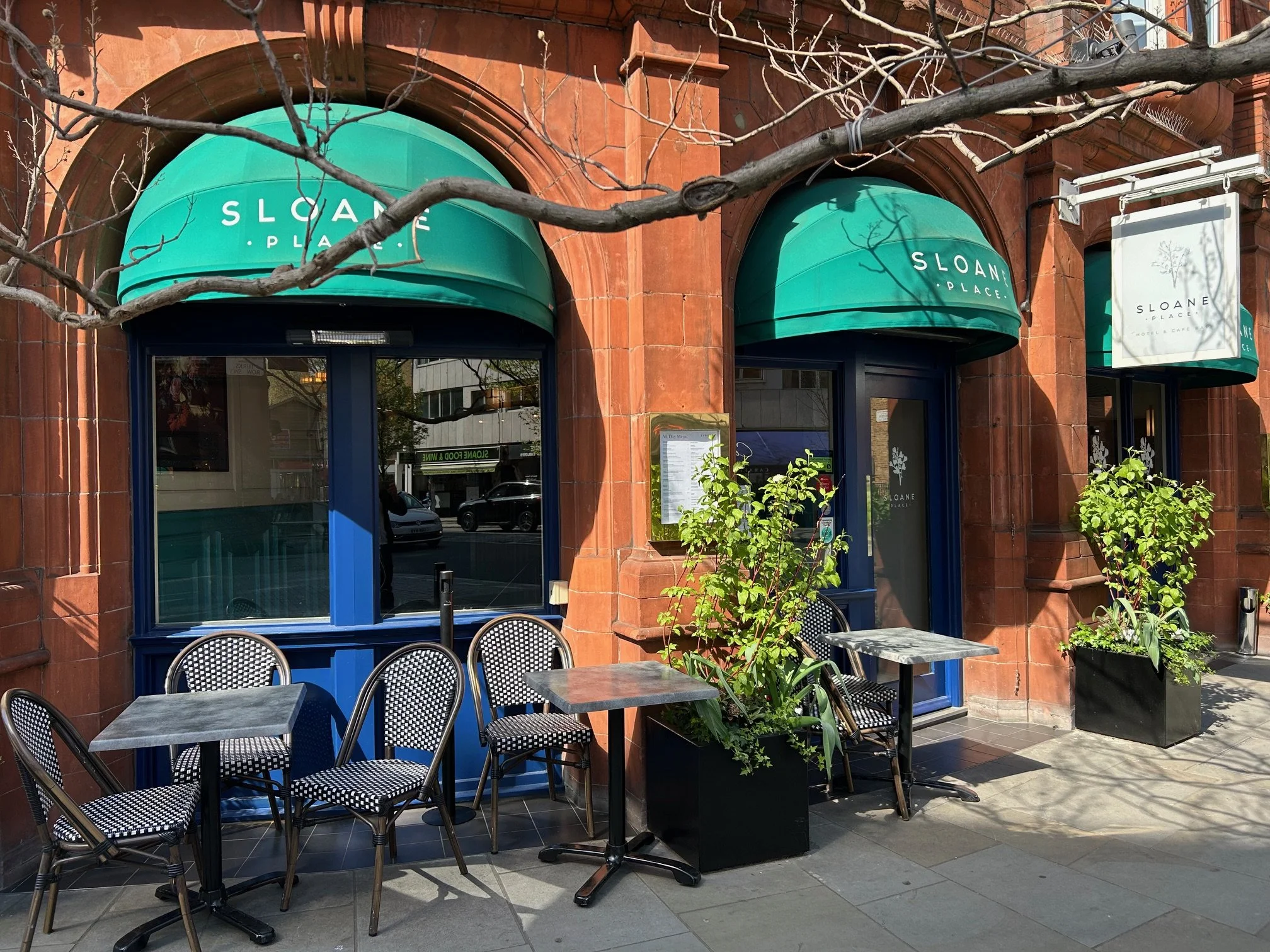 Outdoor seating area in front of a restaurant with blue-framed windows and green awnings that display the name 'SLOANE PLACE'. There are tables and chairs with black and white patterned seats, potted plants, and a leafless tree in the foreground, wit
