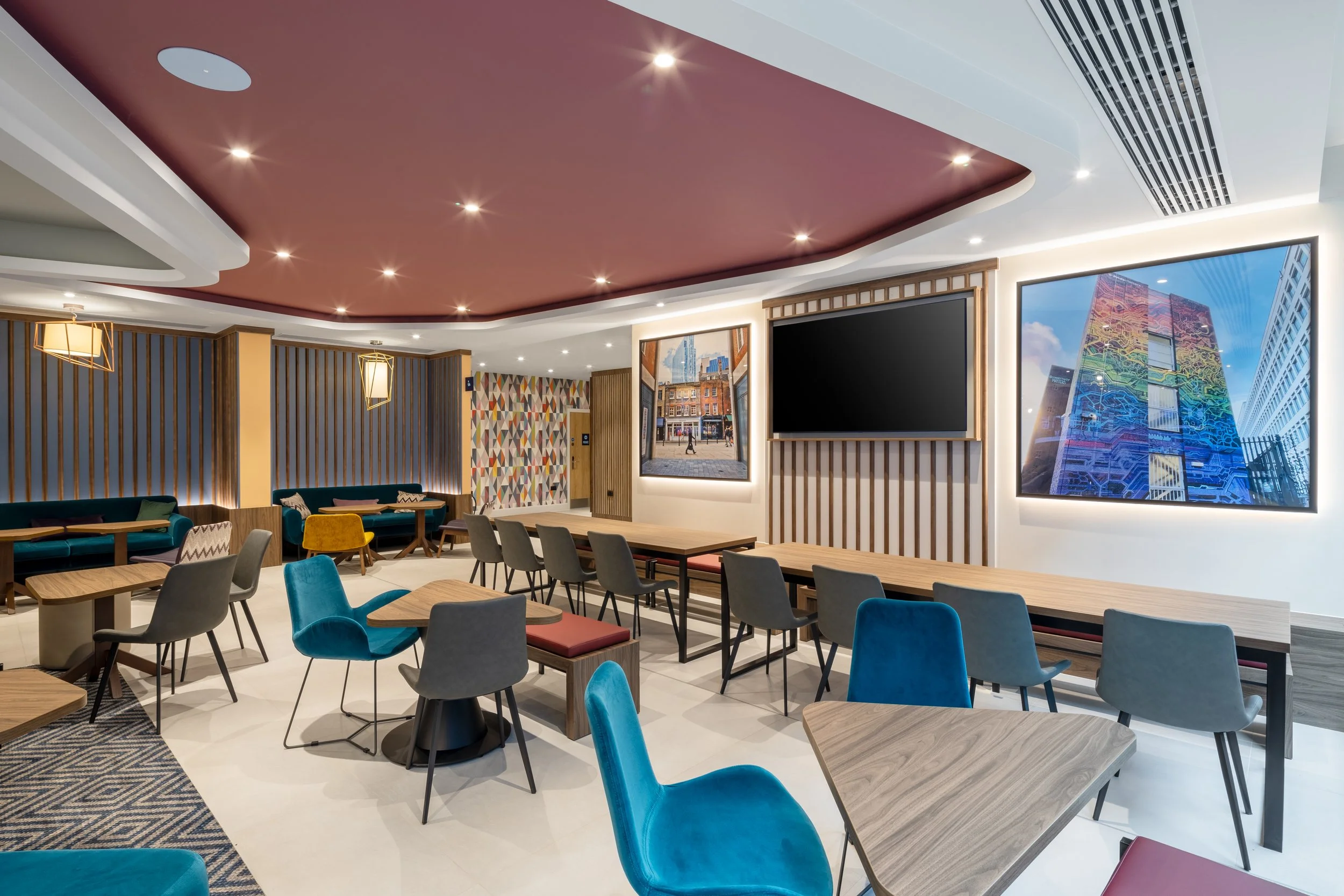 Modern lounge area with colorful chairs, wooden tables, wall-mounted TVs, and large cityscape photographs. Hampton by Hilton Hotel in Old Street
