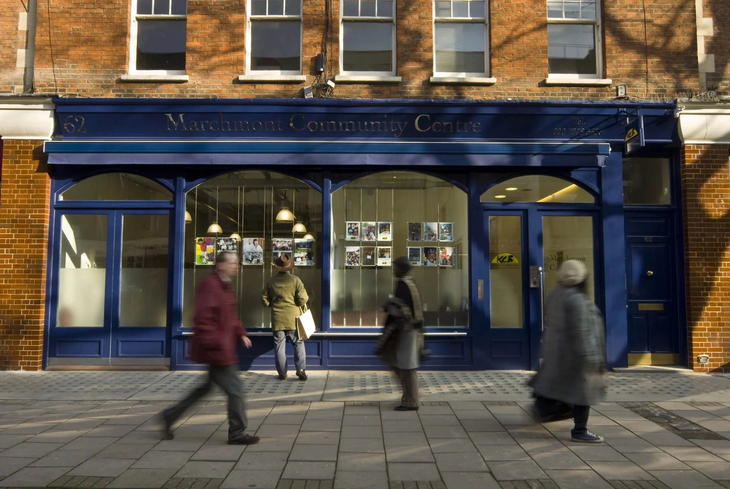 The storefront of the Marchmont Community Centre with a blue facade and large windows, located on a brick building with four upper-floor windows. Several blurred pedestrians are walking past on the sidewalk. Marchmont Community Centre