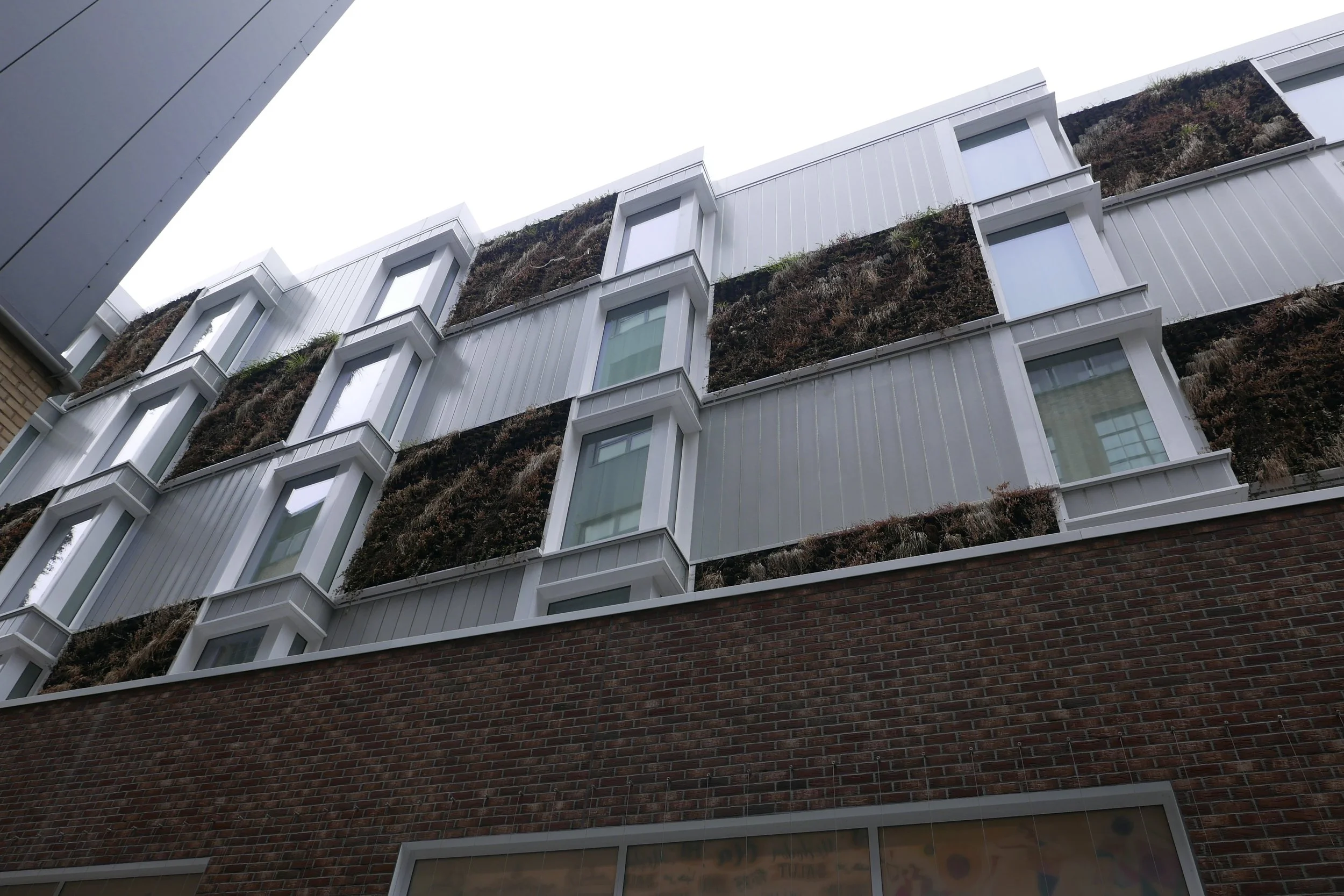 Modern rear façade with white glass panela and brick with protruding white box windows and green plants in the spaces between the building sections. Hampton by Hilton Hotel in Old Street