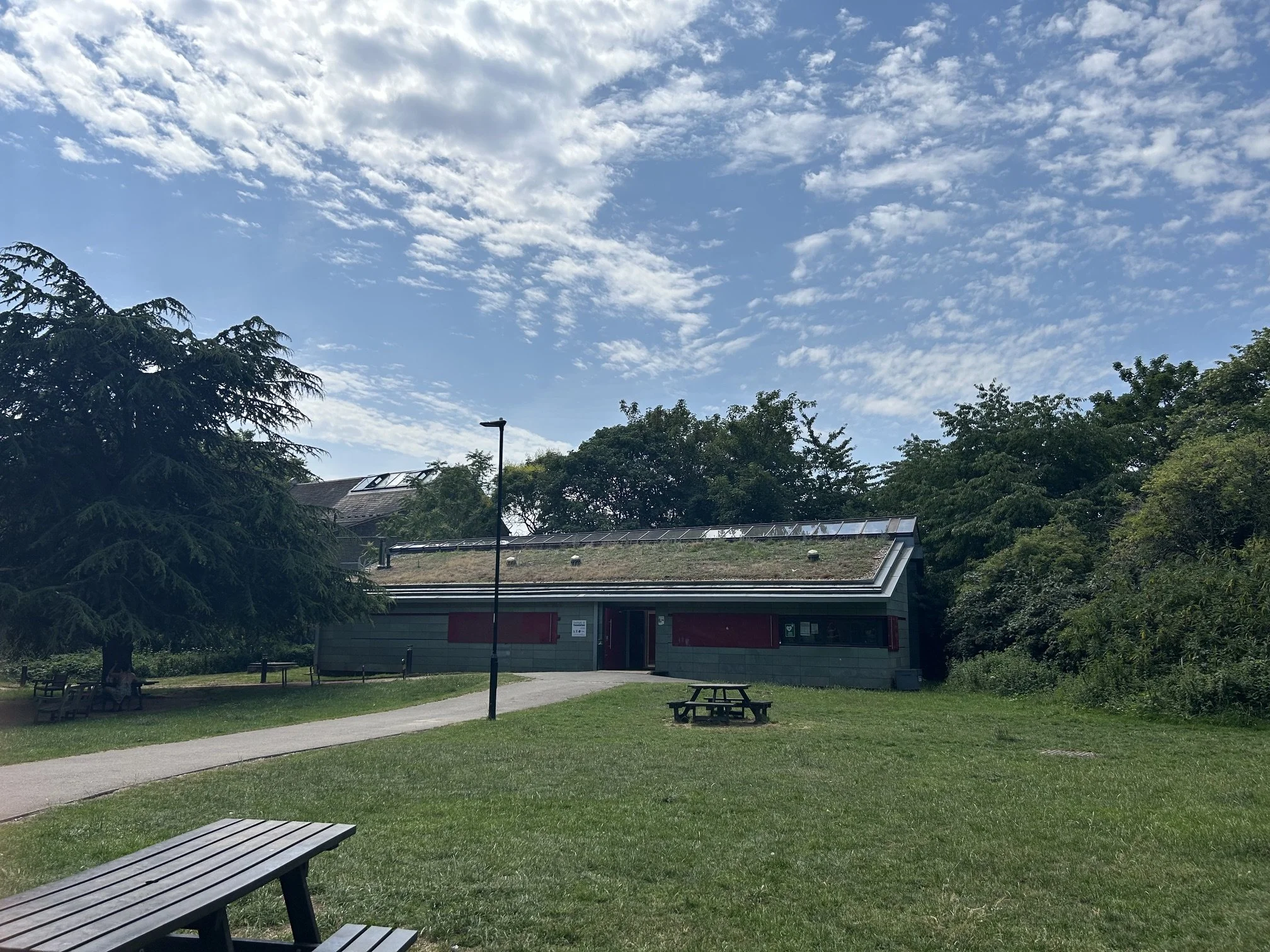 Park views of the new building with green roof , solar panels on the roof, stone cladding and metal shutters, surrounded by trees, a walking path, and a picnic table under .Whittington Park Sport Pavilion