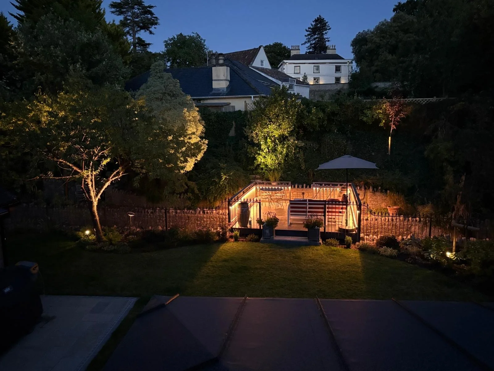 Night view of a backyard patio with outdoor seating, illuminated by garden lights, surrounded by trees and enclosed with a fence.
