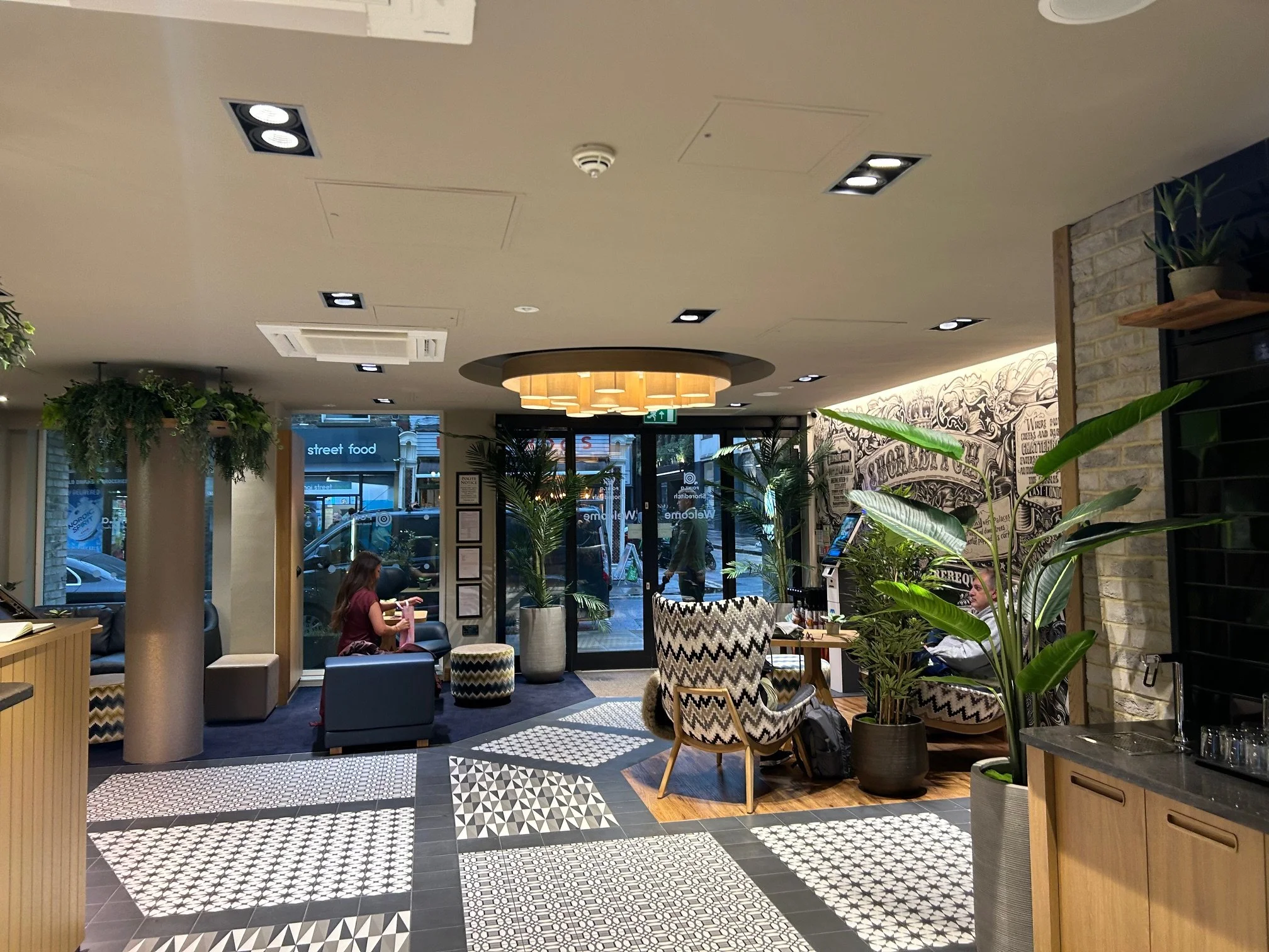 Entrance and reception of the Point A Hotel Shoreditch with modern décor, seating areas with patterned chairs, potted plants, with large windows and street views outside.