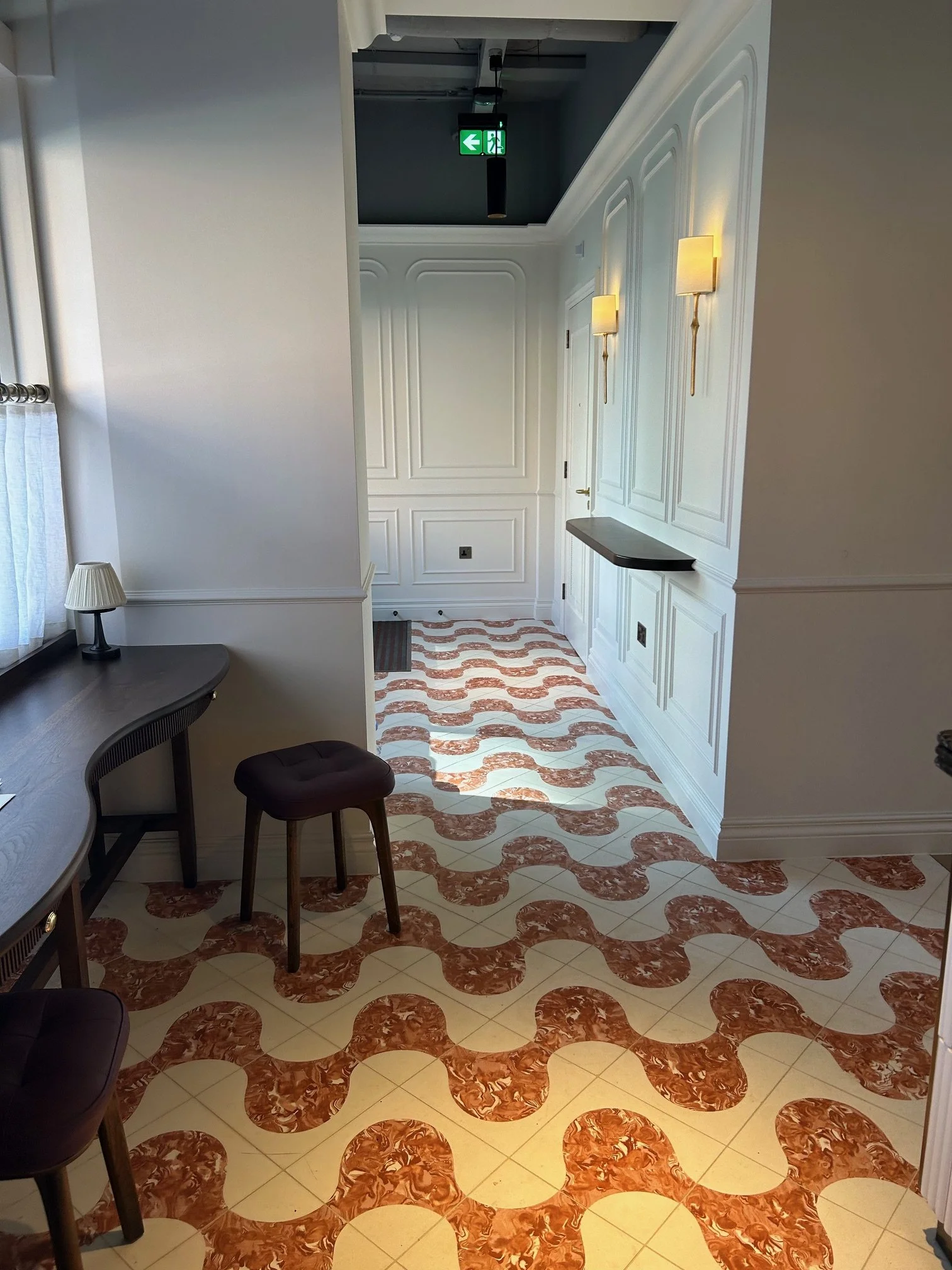 Interior view of a well-lit hallway with white paneled walls, two wall-mounted lamps, patterned floor tiles, a small table with a lamp, a stool, and a green exit sign hanging from the ceiling. The Slone Club Café 1992