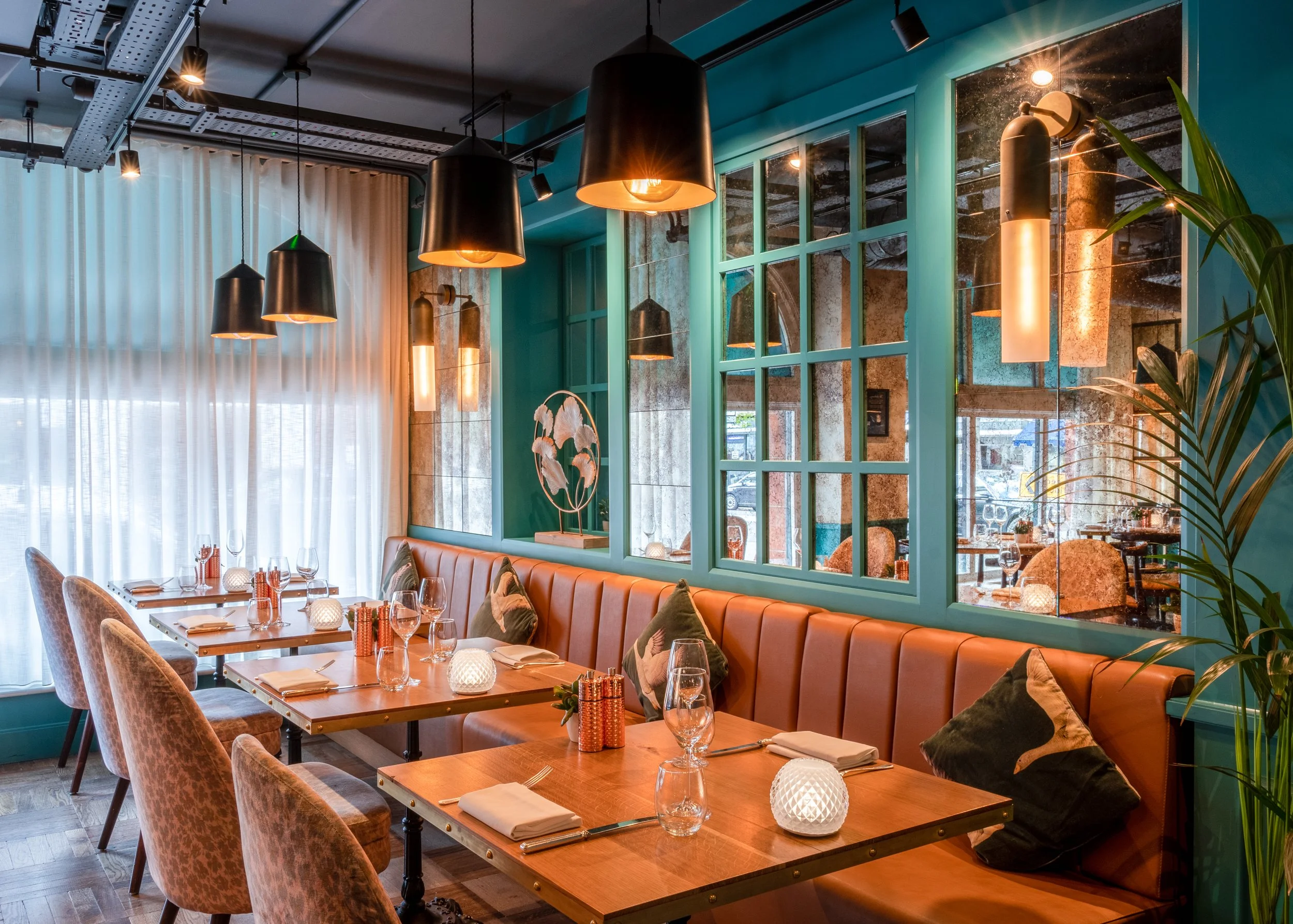 Interior of a stylish restaurant with wooden tables, upholstered chairs, a leather banquette with pillows, modern black pendant lights, decorative lamps, and a large window with sheer curtains. There are some plants and decorative objects on the tabl