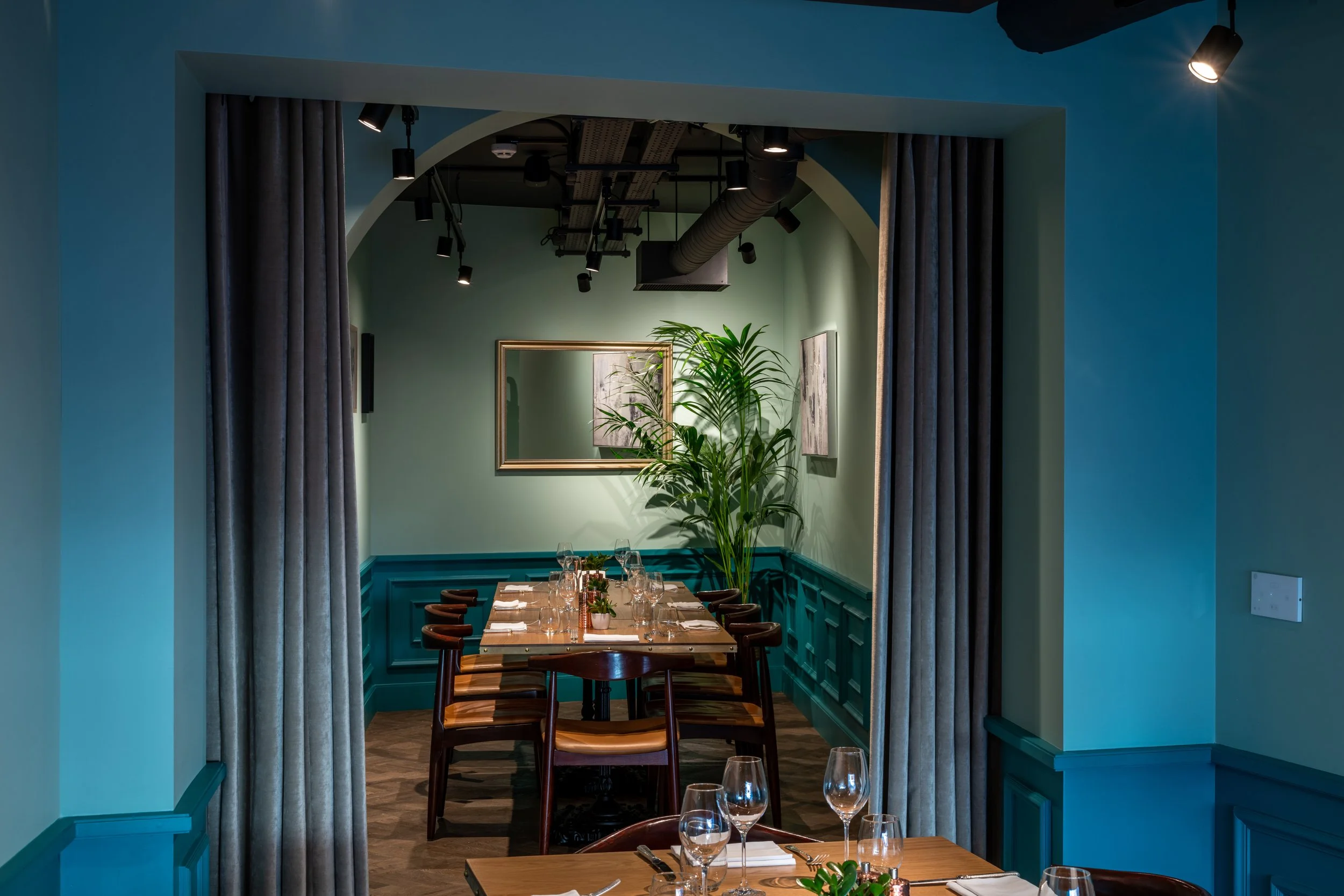 A dining room with teal walls, a wooden table set for a meal, wine glasses, and a centerpiece, viewed through tall curtains. Sloane Place Restaurant