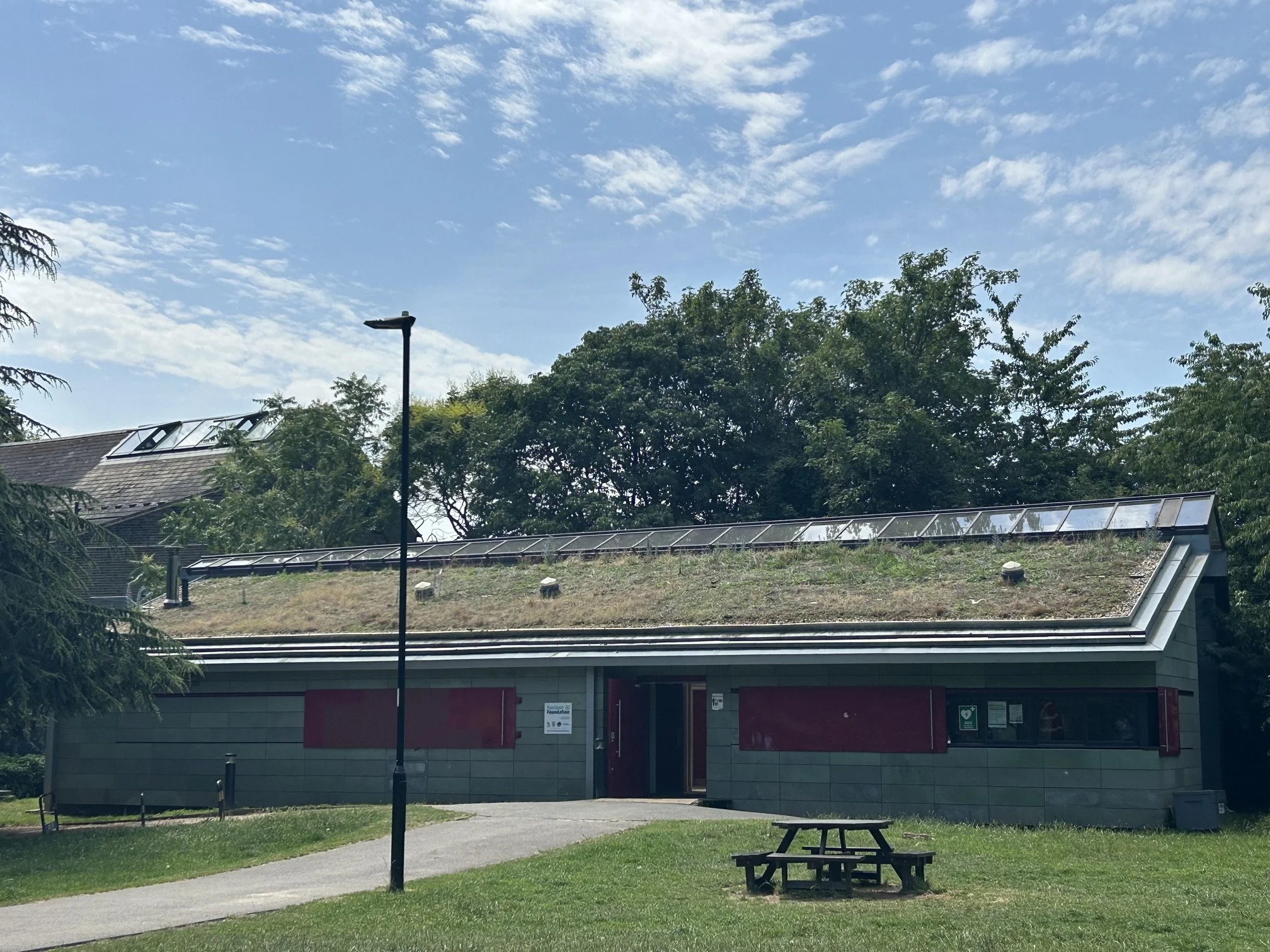 Park views of the new building with green roof , solar panels on the roof, stone cladding and metal shutters, surrounded by trees, a walking path, and a picnic table under .Whittington Park Sport Pavilion