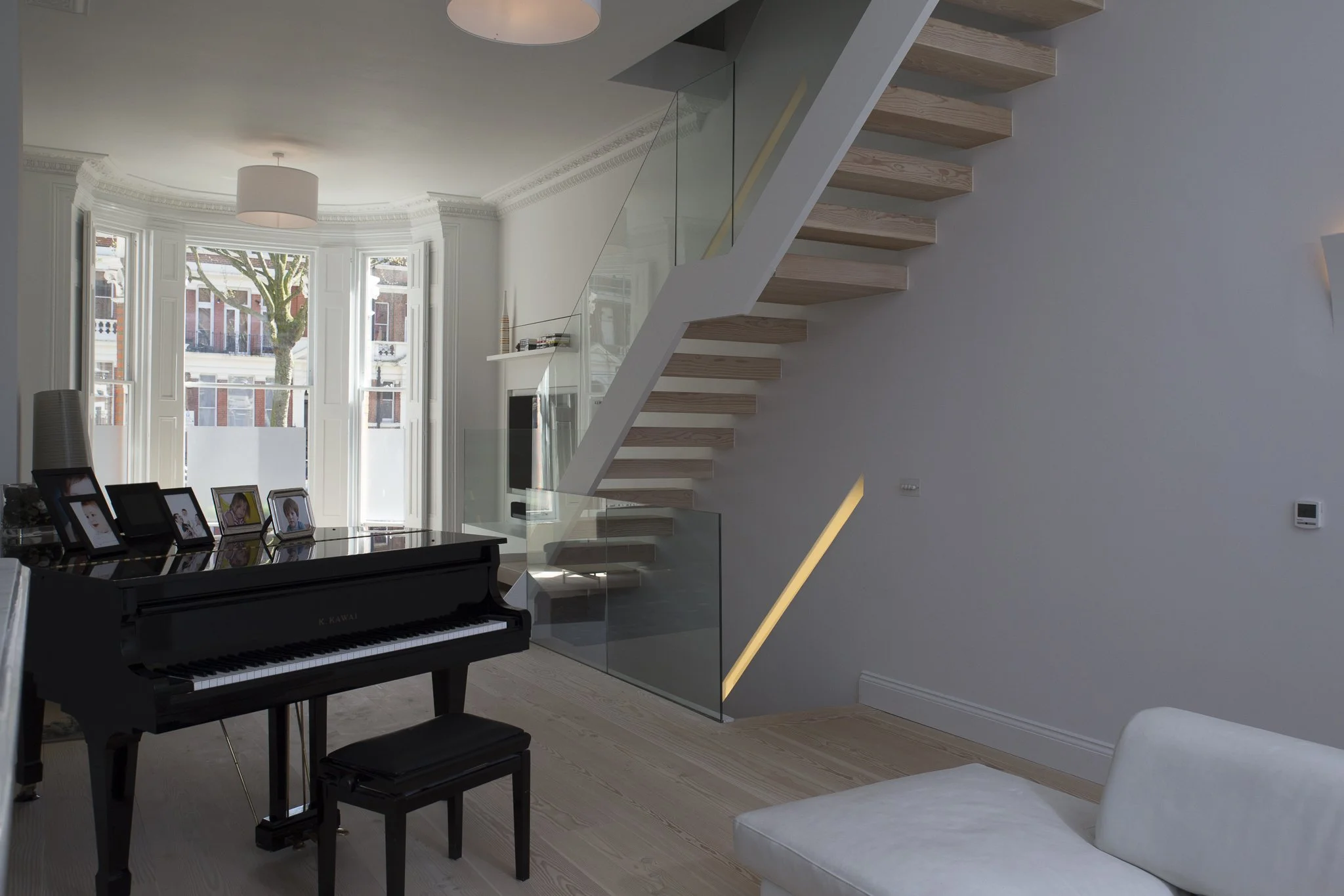 View of the modern living room with a grand piano, large bay windows and the new cantilevered  staircase with wooden steps and glass railing. Sutherland Avenue, Maida Vale