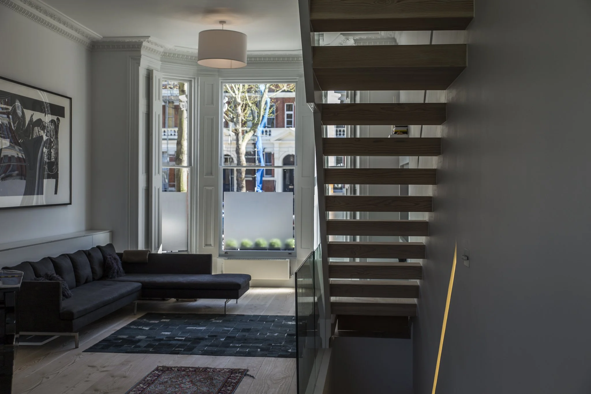 Interior view of a modern living room with large windows, a black sofa, a patterned rug, and a staircase with wooden steps and glass railing, overlooking the street outside. Sutherland Avenue House