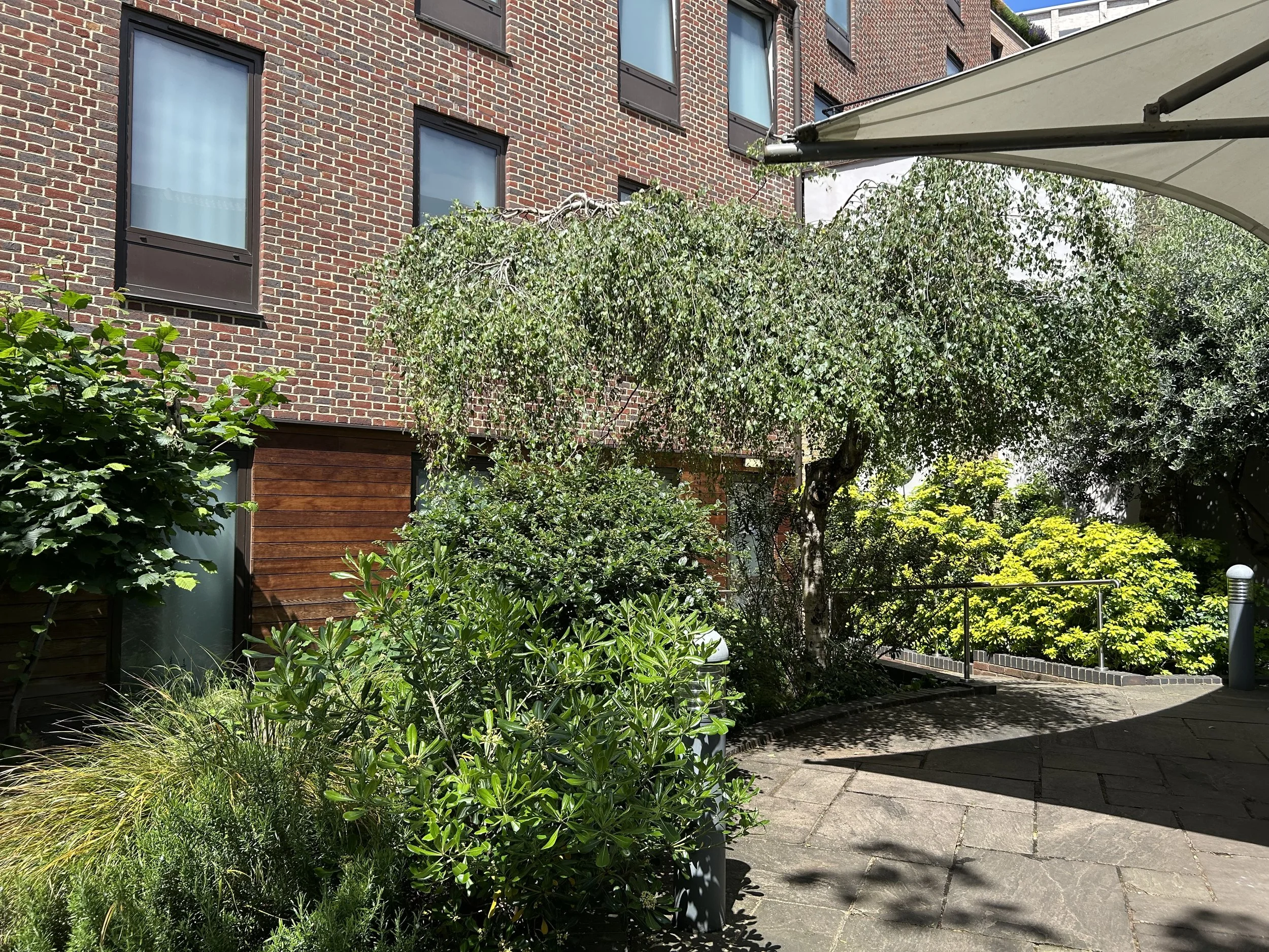 A modern brick extension in the garden of the Point A Hotel Liverpool Street facing lush vegetation, with trees and bushes, and on the right a patio with a large umbrella.