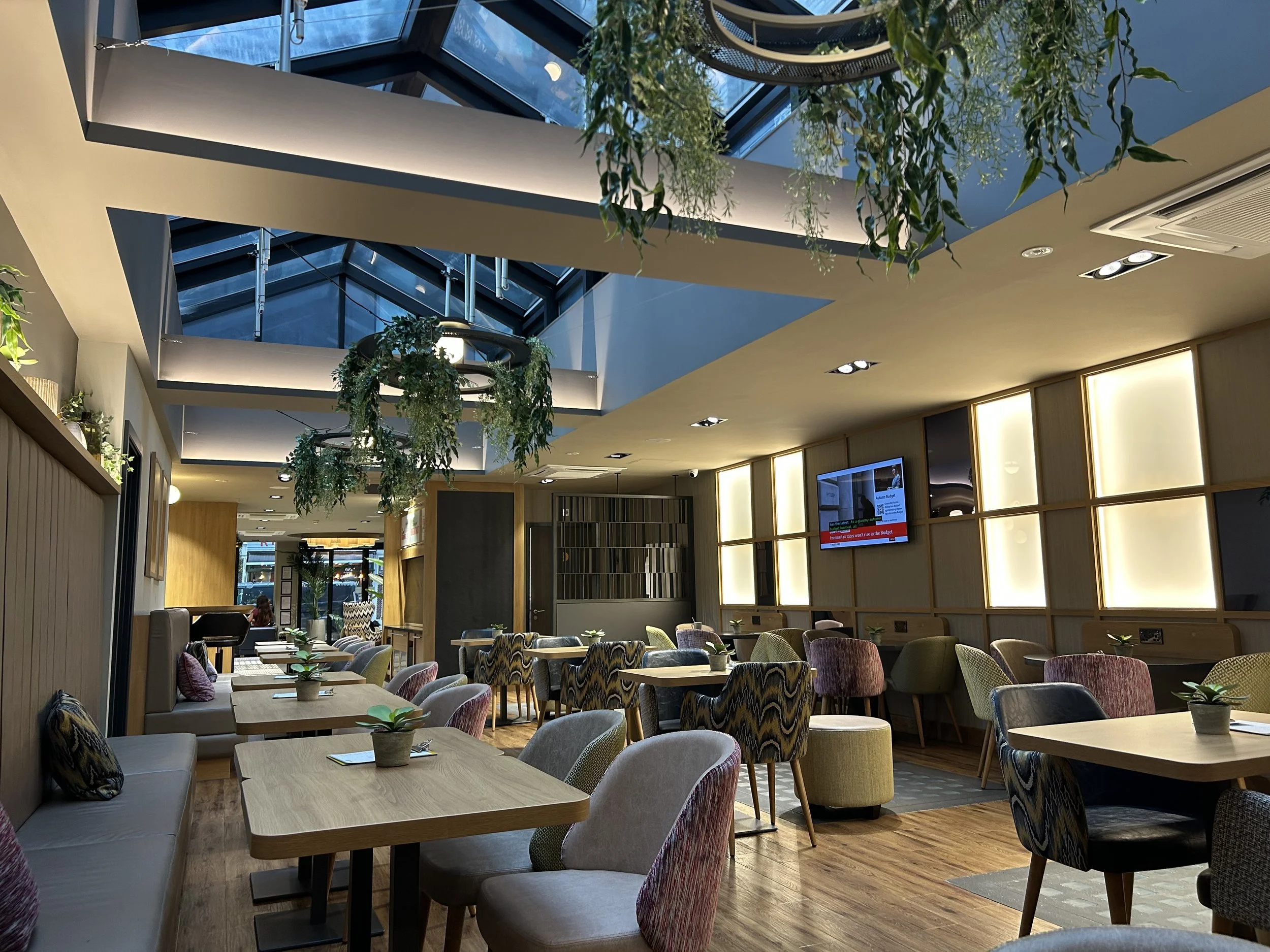 New lounge area of the Point A Hotel Shoreditch with a large skylight with hanging plants and modern design with wooden tables and colourful upholstered chairs.