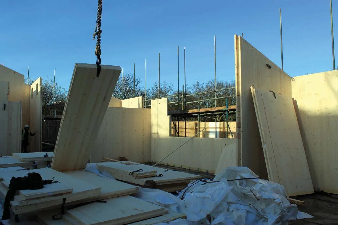 Construction site with wooden walls being assembled, large wood panels on the ground, a crane hook lifting a panel, and scaffolding in the background under a clear blue sky. Whittington Park Sport Pavilion