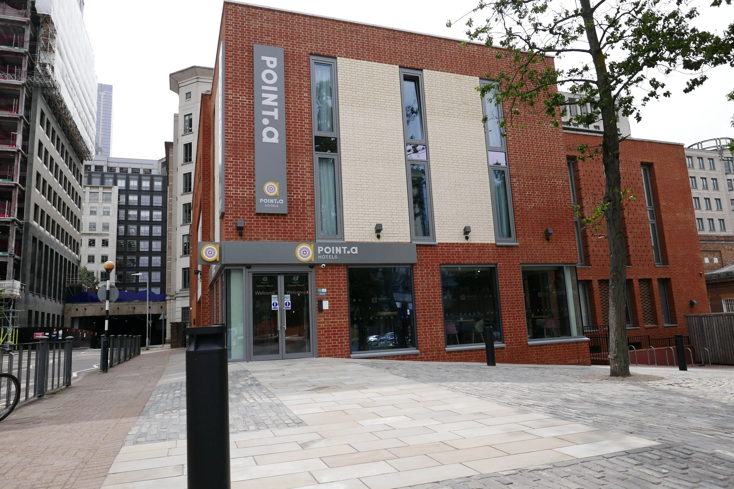 Modern brick hotel building with multiple large windows and a gray sign reading POINT.A HOTELS on a city street, with surrounding high-rise buildings and trees. Point A Hotel Canary Wharf