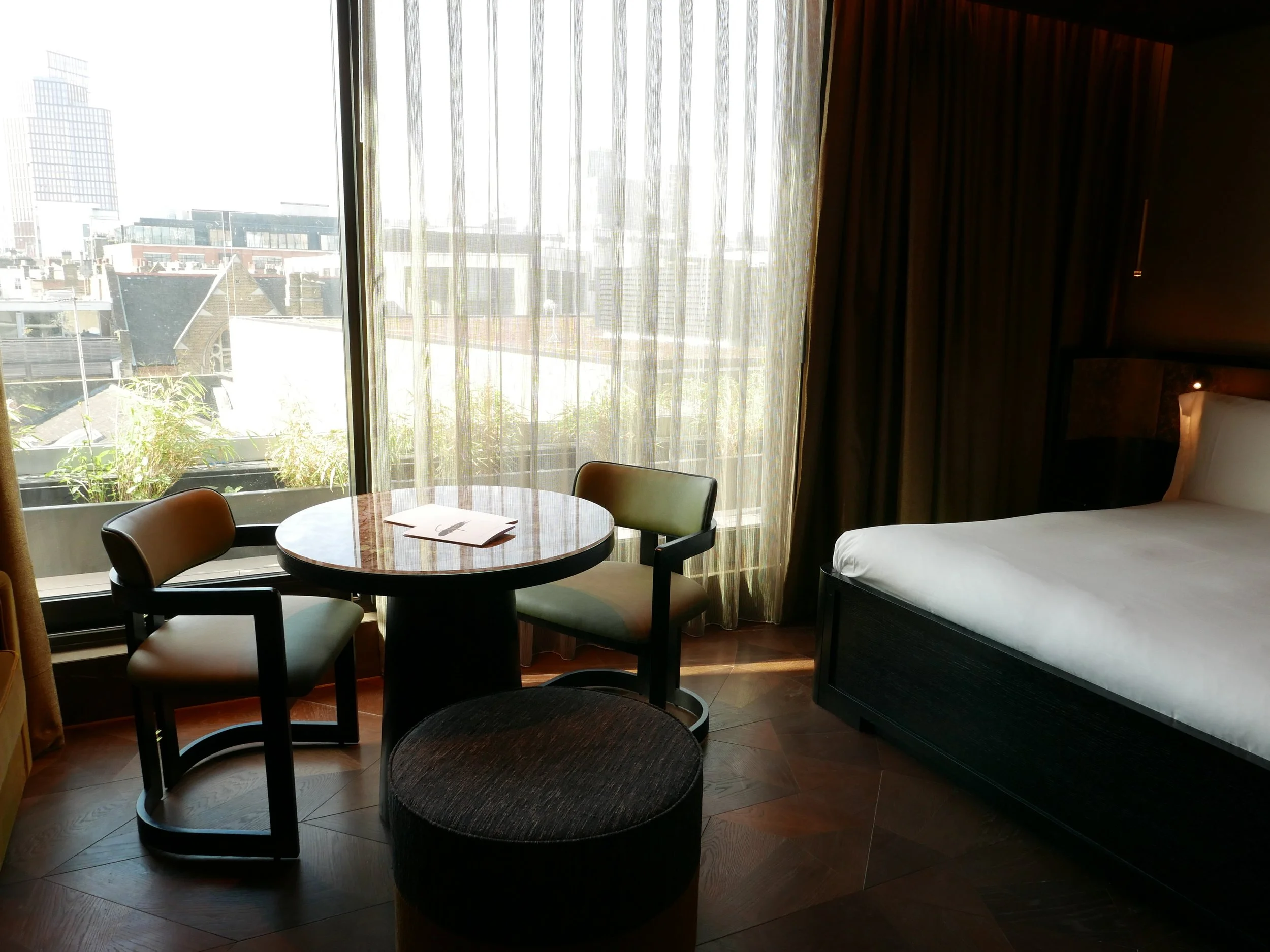 A hotel room with a window view of city buildings, a round dining table with four chairs, a bed with a white pillow, and curtains partially covering the window.
