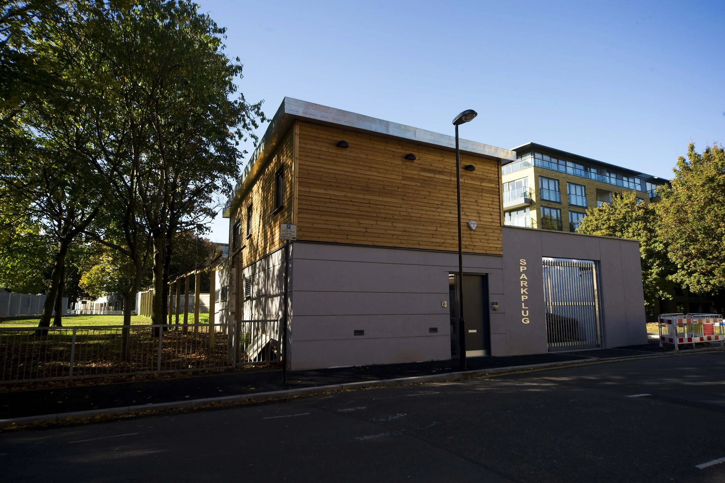 Street view of the modern building with grey render wall at ground floor and wooden cladding on the first floor.  SPARK PLUG" metal letters next to the large gate, situated along a street. Sparkplug