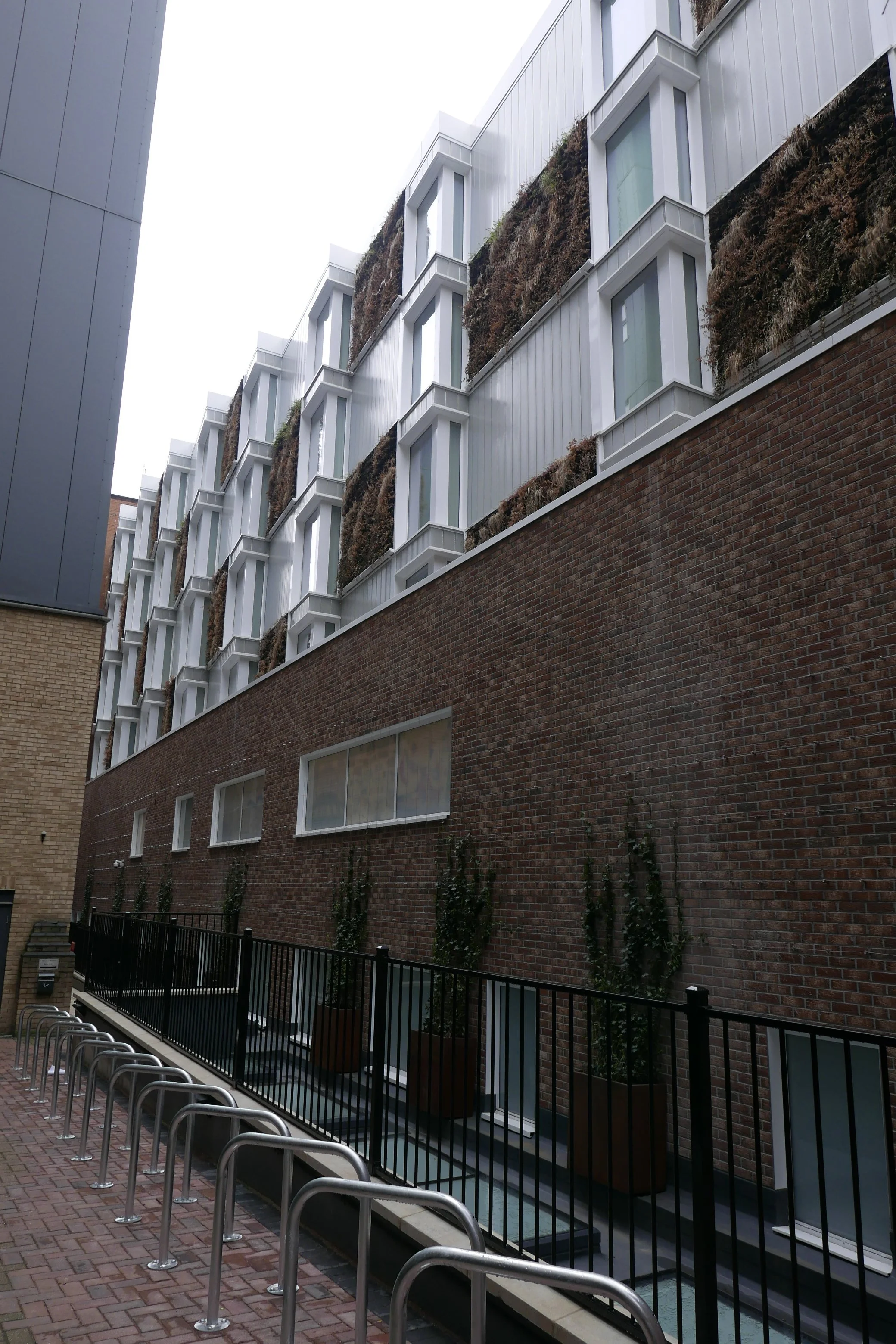 Modern rear façade with multiple story levels, brick lower wall, glass windows with white framing, and vertical gardens with plants in wooden planters, of the new Hampton by Hilton Hotel in Old Street