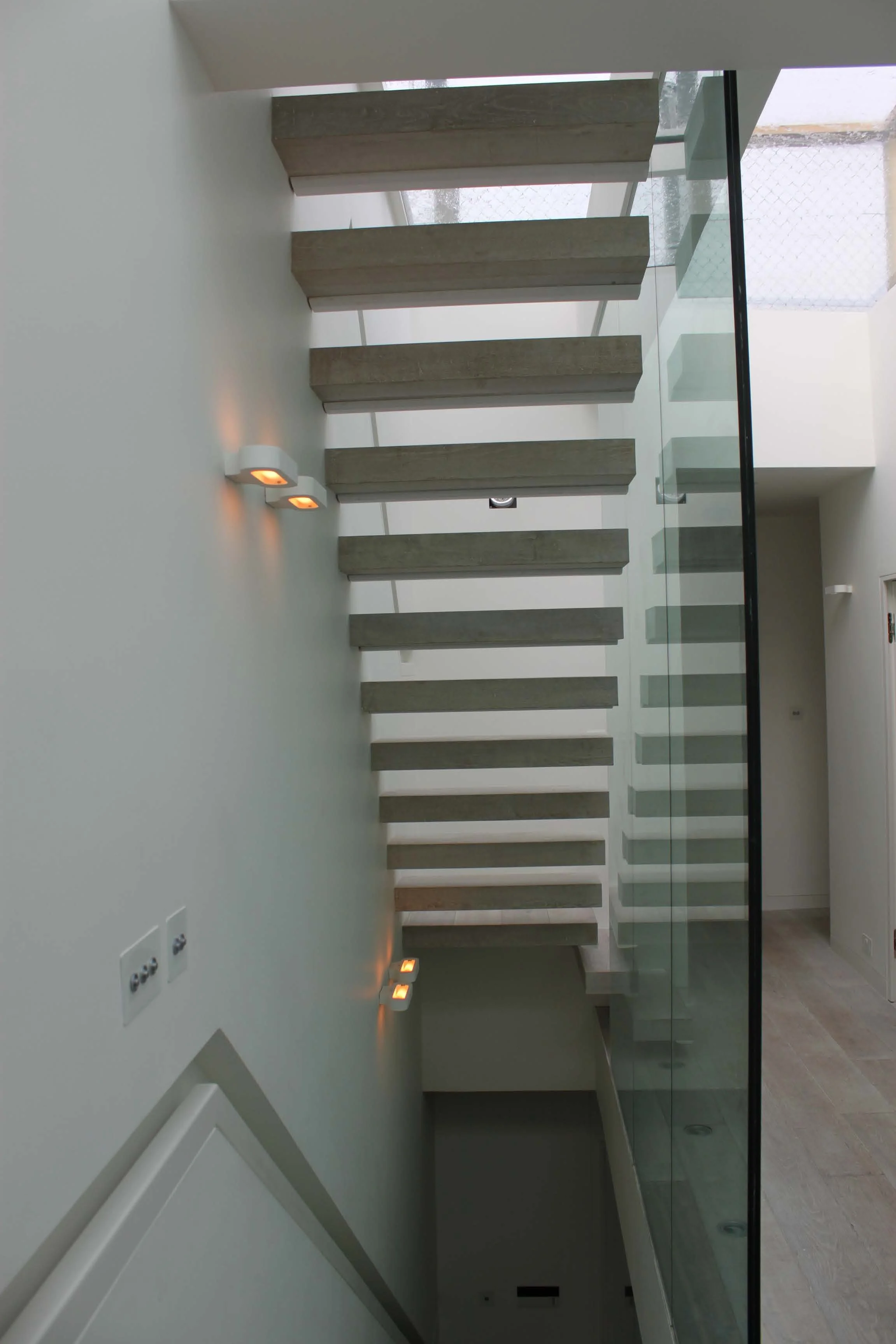 Modern interior with a staircase featuring wooden steps and a glass railing, white walls, and small wall-mounted lights.. South Kensington House