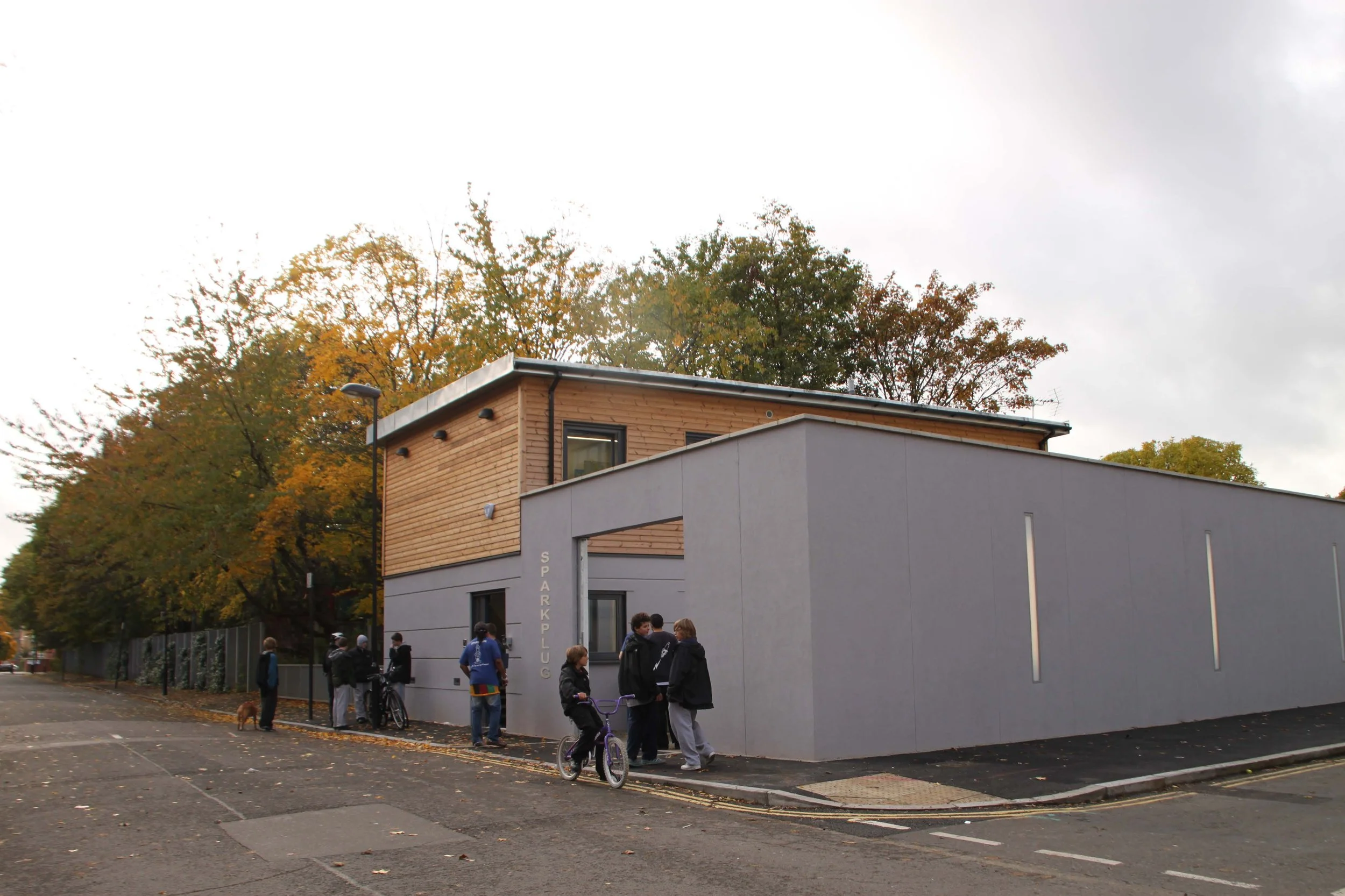 External corner view of the new building with the high grey rendered wall with recessed lighting around the courtyard and view of the modern building with wooden cladding.  "SPARK PLUG" metal letters next to the large gate. Sparkplug