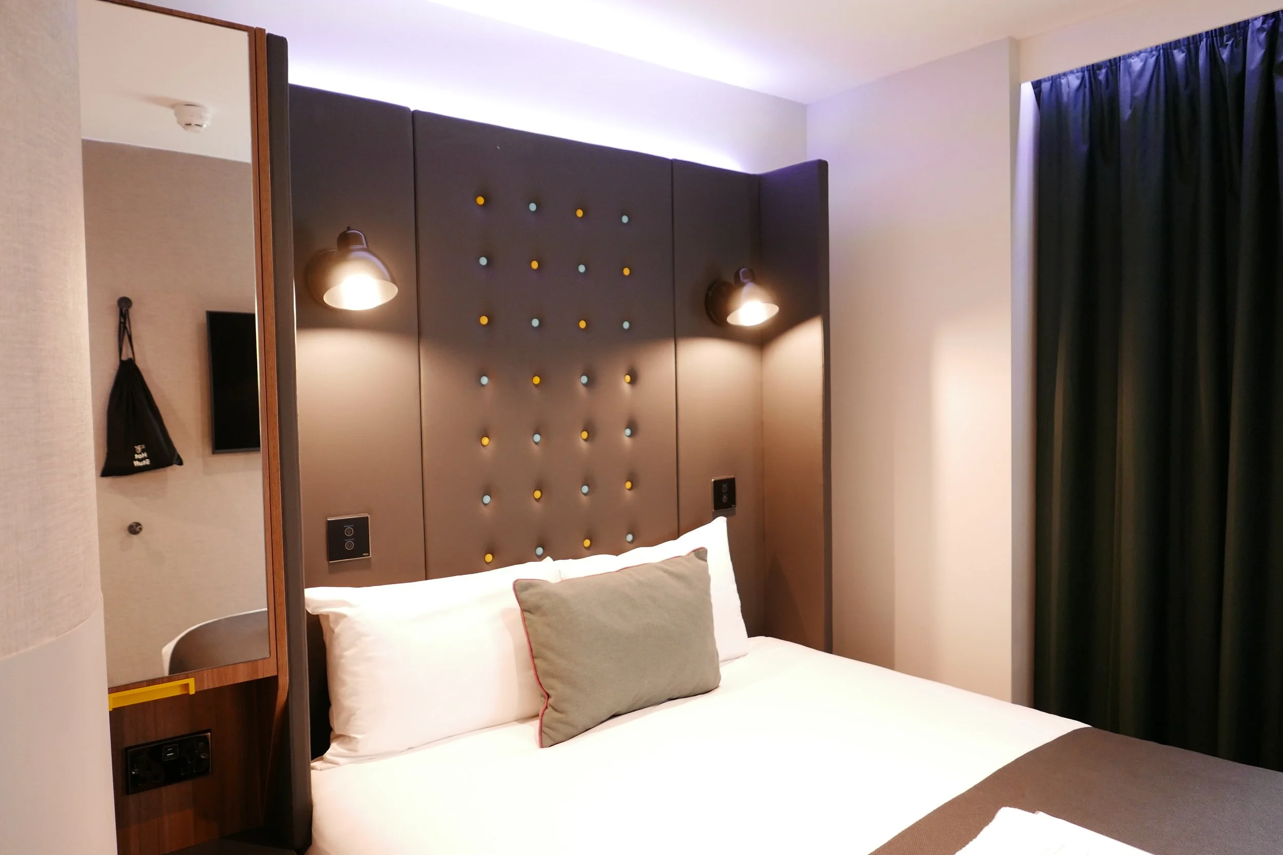 Modern hotel bedroom with a large bed, decorative headboard with coloured lights, wall-mounted lamps, a mirror of the Point A Hotel Shoreditch