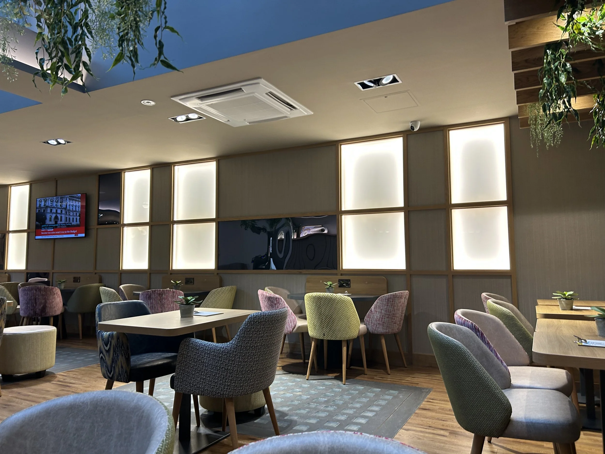New lounge area of the Point A Hotel Shoreditch with a large skylight with hanging plants and modern design with wooden tables and colourful upholstered chairs.
