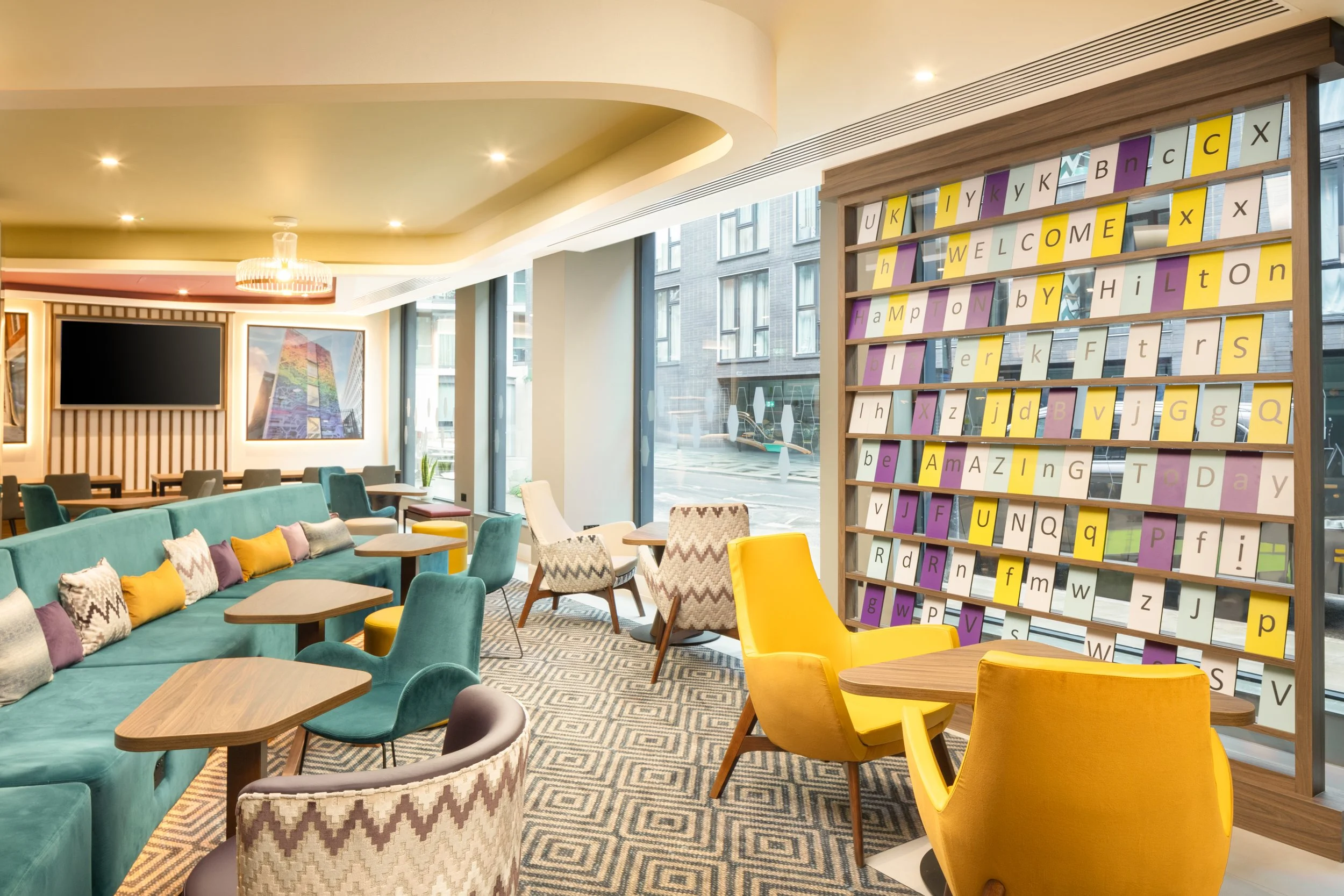 Interior of a modern lounge or hotel lobby with colorful furniture, a large window, and a decorative book display spelling 'Welcome' and other words. Hampton by Hilton Hotel in Old Street
