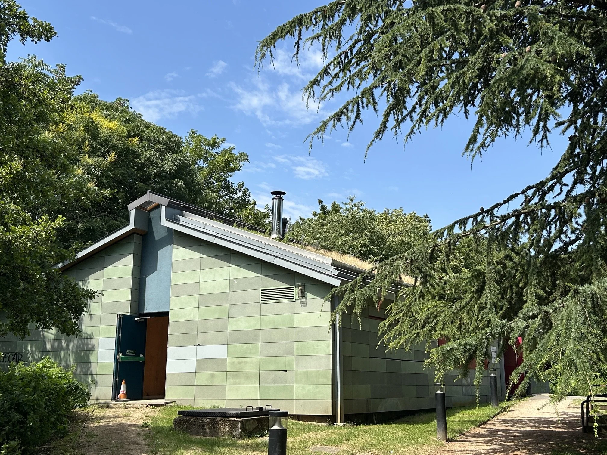 Detail of the modern building with green tiled walls and a doorway, surrounded by trees and greenery, under a bright blue sky with some clouds. Whittington Park Sport Pavilion