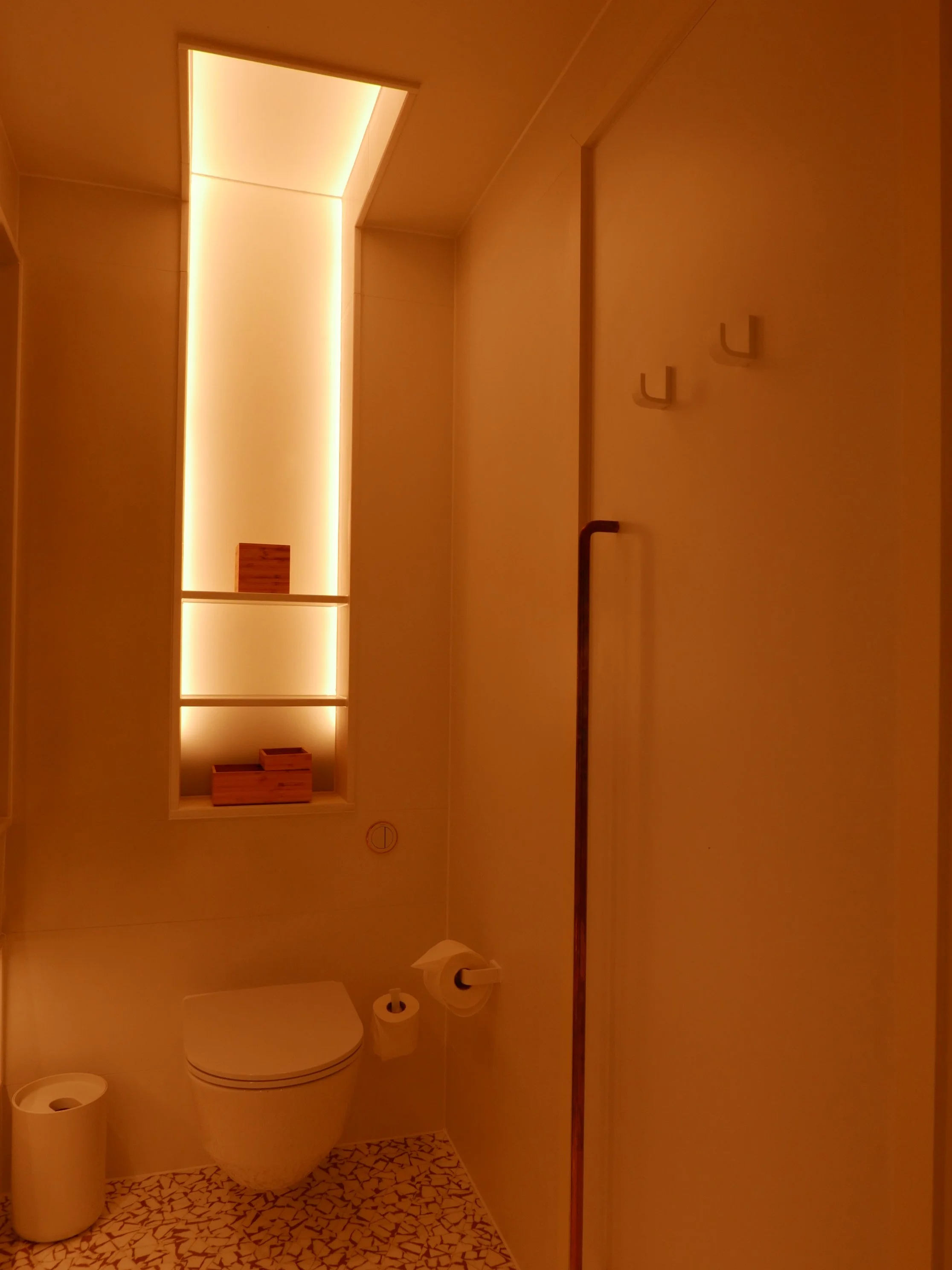 A compact bathroom with warm lighting, featuring a wall-mounted toilet, a toilet paper holder, and a narrow wall-mounted shelf with wooden boxes, illuminated by vertical LED lighting. Nobu Shoreditch roof extension