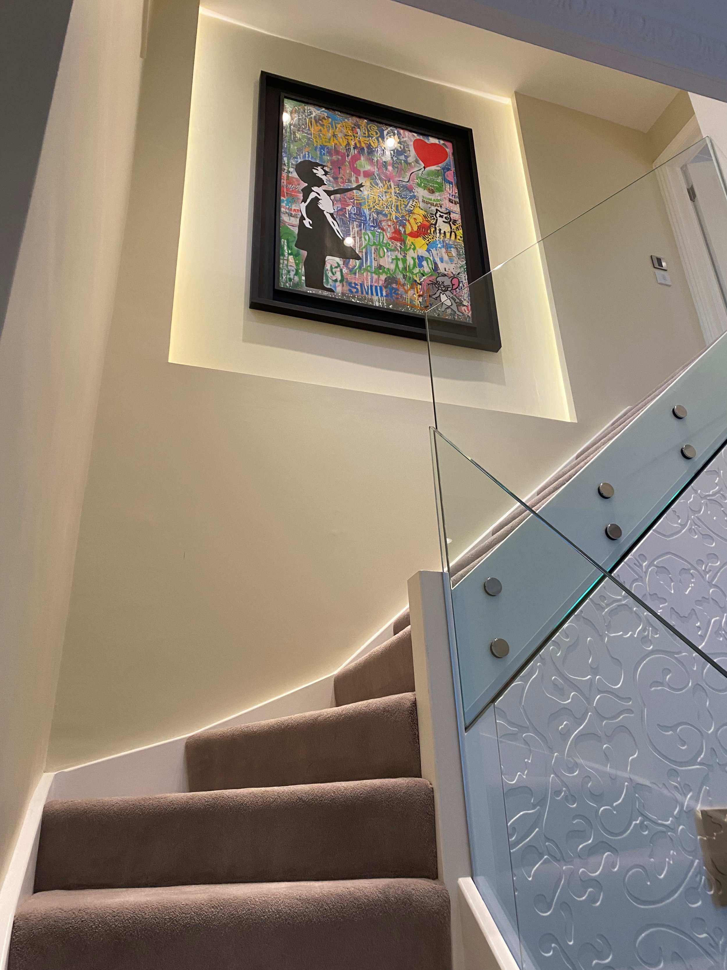 Indoor staircase with beige carpeted stairs and a white carved railing on the right side. There is a framed artwork with colorful graffiti-style design hanging on the wall above the stairs. The wall behind the artwork is illuminated with warm lightin