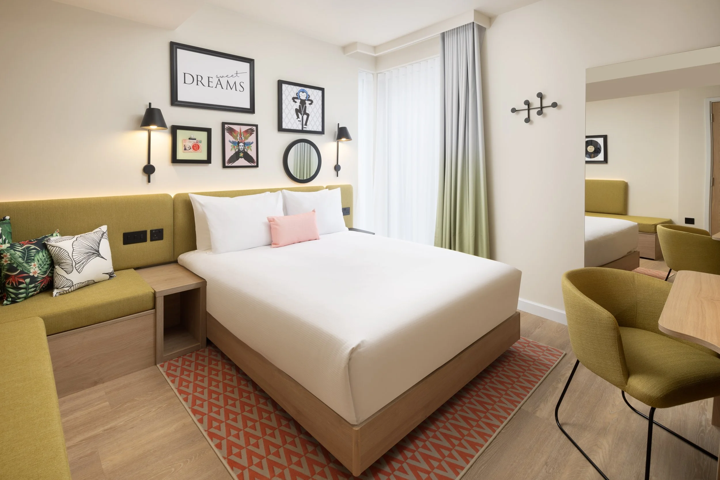 A modern hotel room with a bed, yellow-green headboard, framed artwork, wall lamps, and a window with curtains. Hampton by Hilton Hotel in Old Street