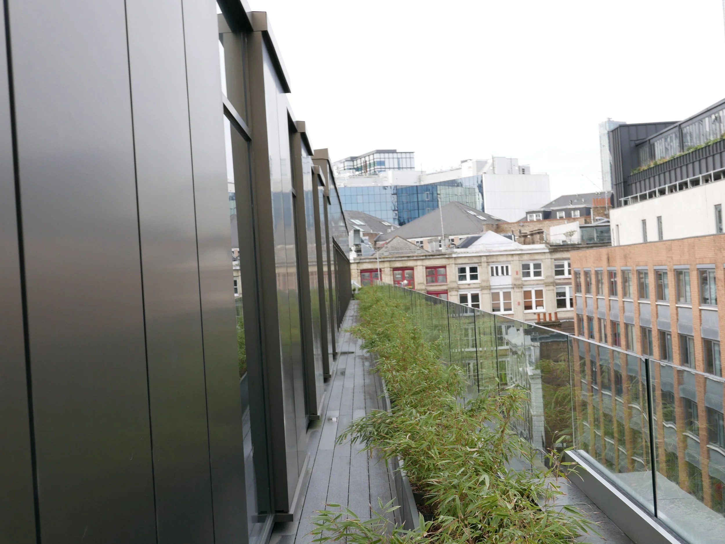 Top floor bedroom modern balcony with glass railing overlooking city buildings, some with reflective glass, under an overcast sky. Nobu Shoreditch roof extension