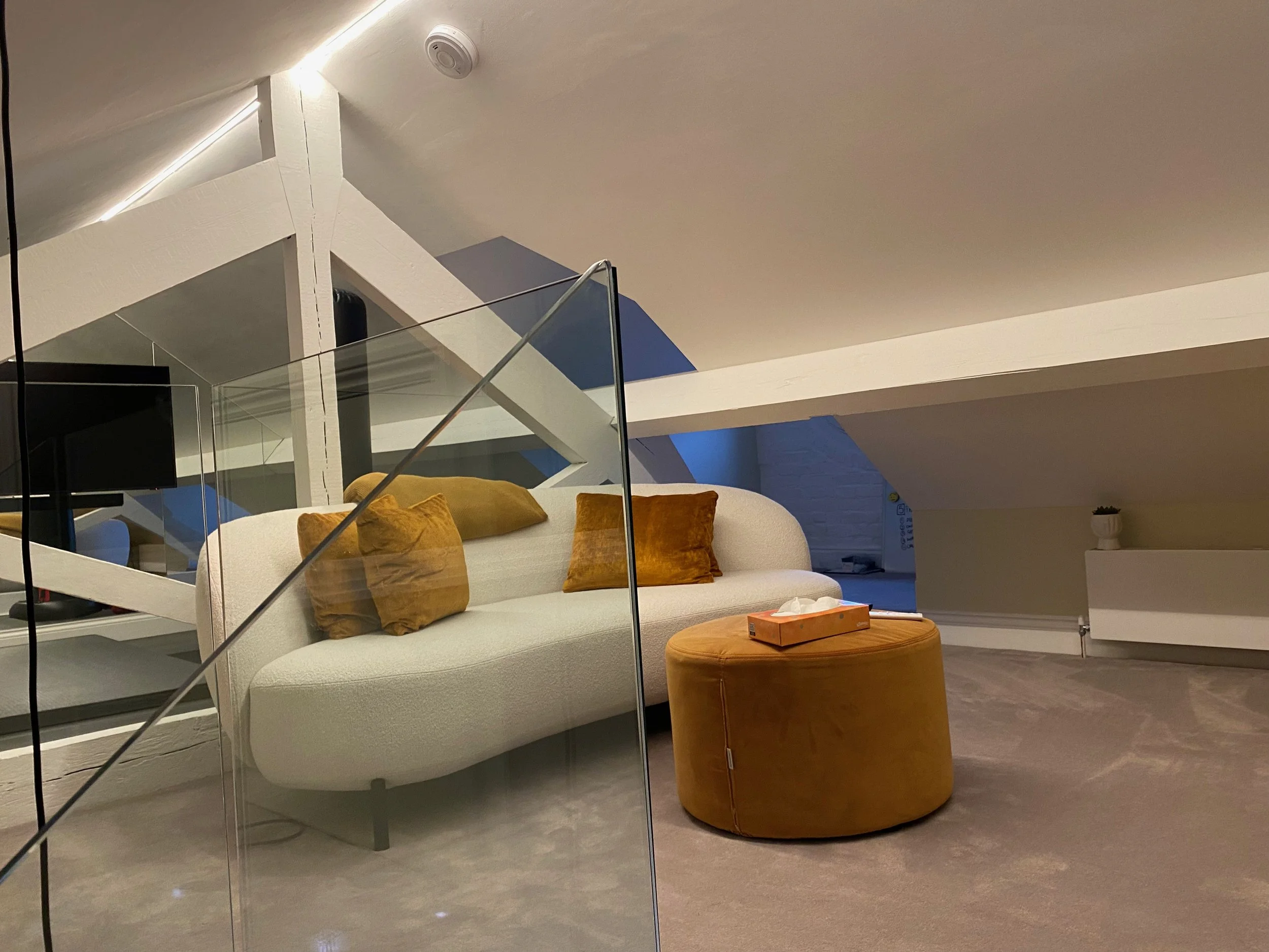 Modern living space in roof void with white sofa, mustard and brown cushions, round mustard ottoman, glass railing, staircase, and minimal decor. Pont Street Knightsbridge 
