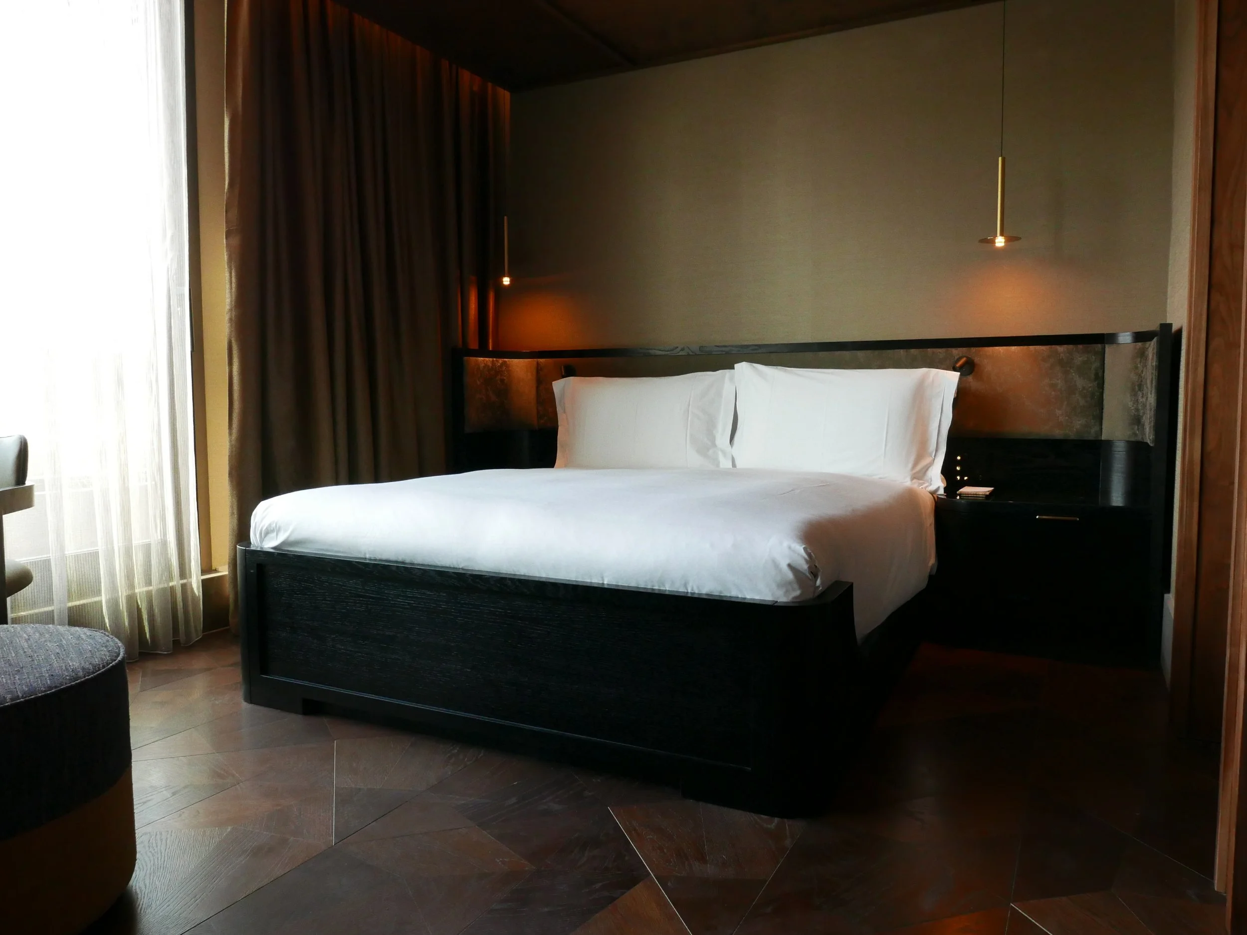 A modern hotel room with a bed, dark wooden headboard, gold pendant lights, curtains, and a small bedside table.